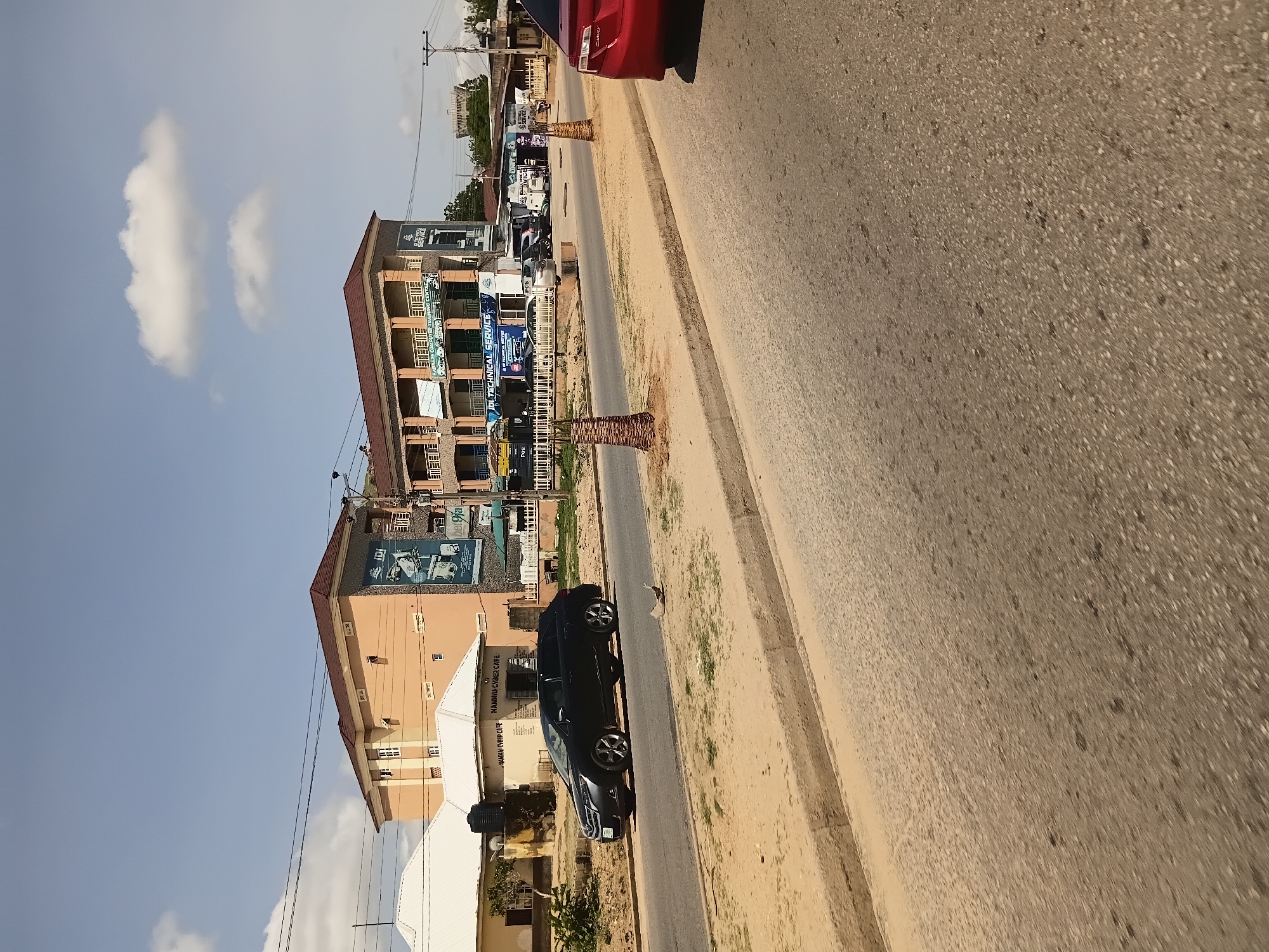 The image depicts a street scene in Jalingo, Nigeria. The prominent structures include a multi-story building with advertising banners, likely housing businesses, and a smaller single-story building with a white roof. A black car is parked on the side of the road, and a red car is partially visible in the foreground. The road is paved, and there is a dirt or sandy median with some vegetation. The sky is blue with scattered white clouds. Power lines are visible overhead, indicating urban infrastructure. The buildings appear to be constructed with concrete and painted in various colors. One banner on the multi-story building displays "ADI TECHNICAL SERVICE" and "IDI COMPUTER TRAINING CENTRE." The overall impression is that of a commercial area on a sunny day.