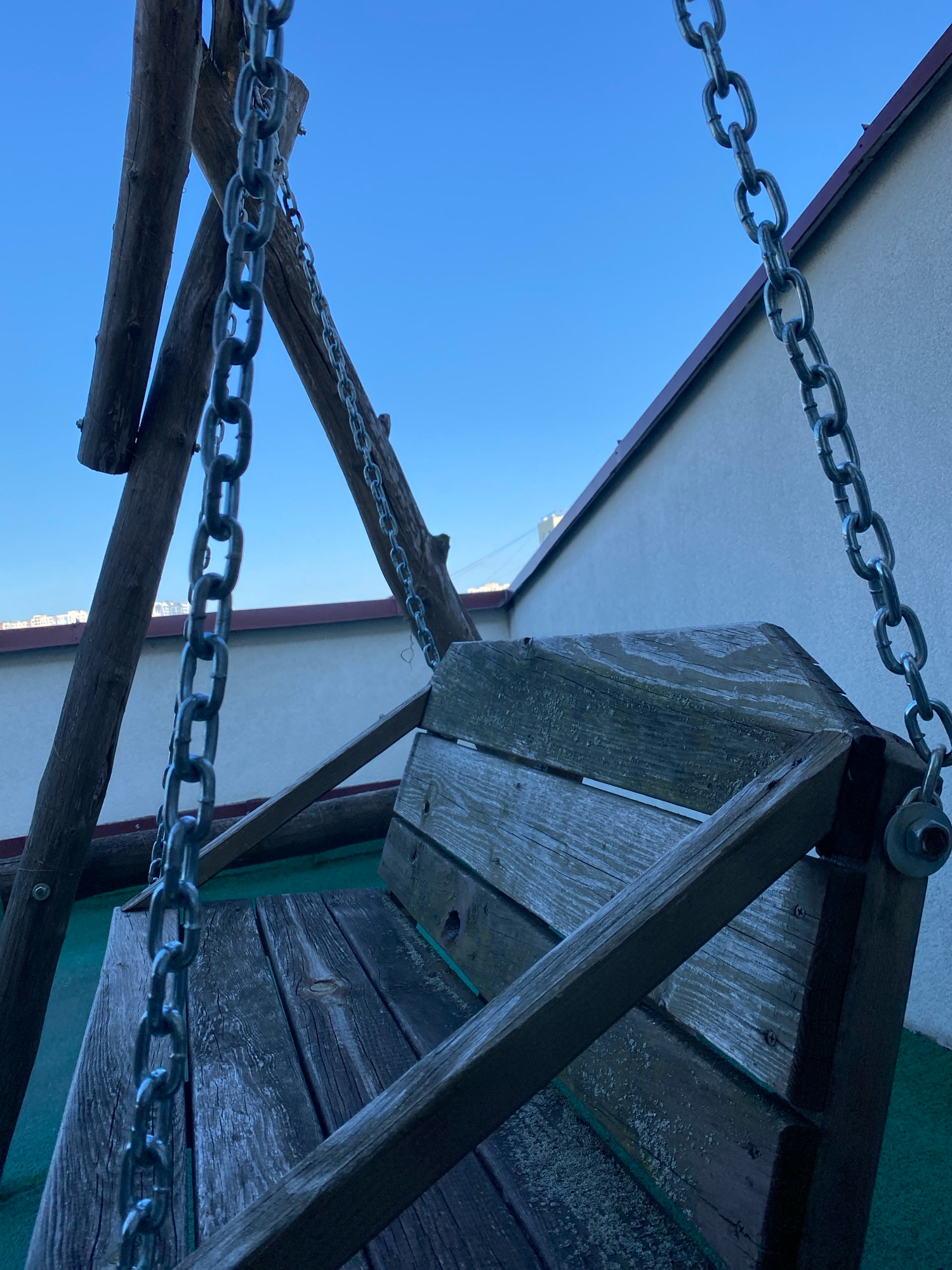 The image shows a close-up of a rustic wooden swing set with metal chains, against a clear blue sky and a light-colored building with a dark roofline. The swing seat is made of weathered wooden planks, and the frame appears to be constructed from rough-hewn logs. The scene is outdoors, likely on a rooftop or balcony given the building in the background. No people are visible. The camera angle is low, looking upwards towards the sky.