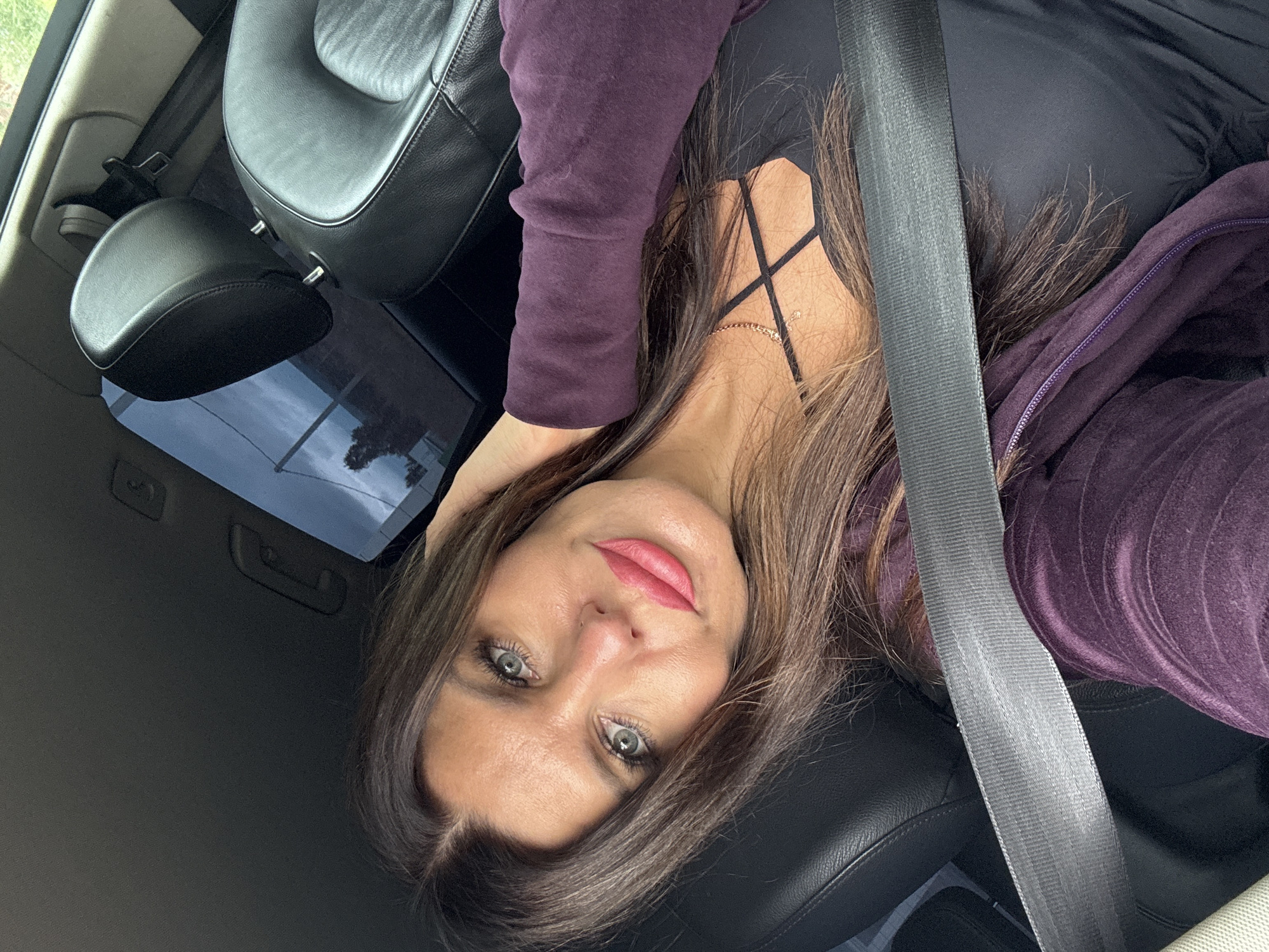 A woman with long brown hair and blue eyes is seen from the chest up, taking a selfie in a car. She is wearing a purple zip-up hoodie over a black tank top and a gold necklace. Her left hand is resting on her head. The background shows a car interior with black leather seats and a seatbelt across her chest. The exterior visible through the car window appears to be a cloudy sky and some trees. There are no identifiable landmarks or indicators of the specific location within Kvarsa, Russia.