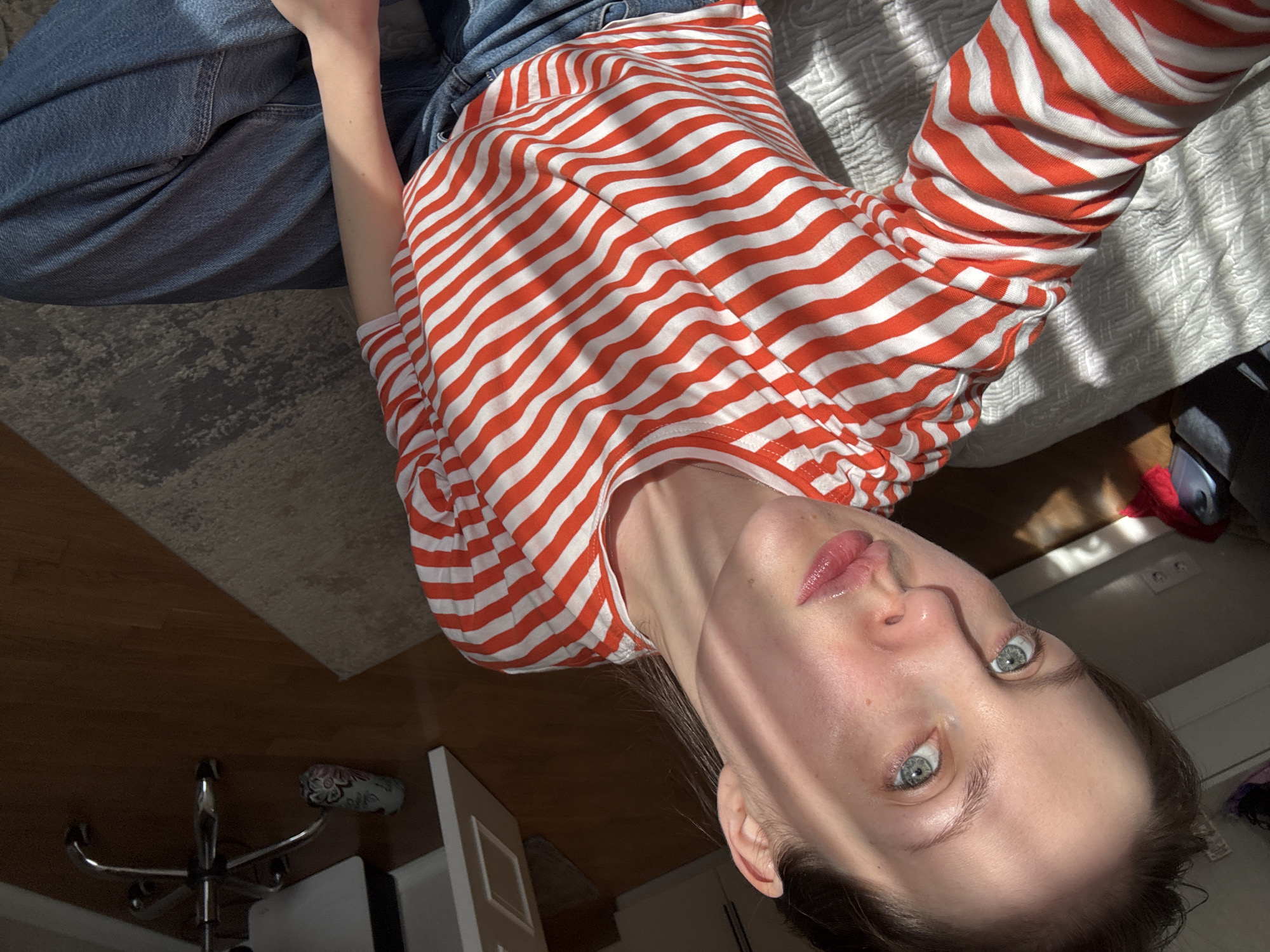 A low-angle, indoor shot depicts a person wearing a red and white striped long-sleeved shirt and blue jeans. The person is lying on a bed, with a portion of a grey patterned rug visible on the left. A desk chair is visible in the lower left corner. The wall and a partially visible door are in the lower part of the image. The environment suggests a bedroom. There is no information to confirm the location is Moscow, Russia.