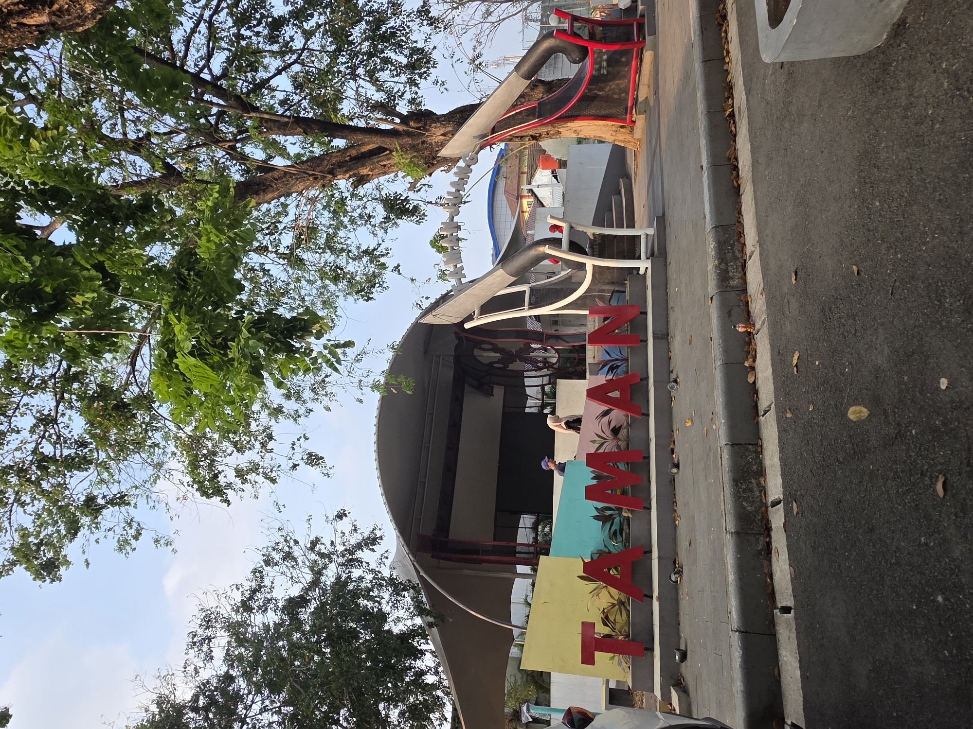 This image captures an outdoor scene in Tambun, Indonesia. In the foreground, large red letters spell out "TAMAN," a common word for park or garden in Indonesian. These letters are positioned on a raised, multi-colored platform featuring yellow, blue, and pink sections, with stylized plants depicted behind them.
Behind the "TAMAN" sign, a modern, curved roof structure with a light-colored fabric canopy extends over an open-air area. Within this covered space, two individuals are visible: one wearing a blue cap and a dark shirt, and another with a light-colored hijab and a dark outfit. A large, intricate metal sculpture resembling a spinning wheel or a circular frame is partially visible within this area, and to its right, a red and black abstract sculpture, possibly representing a large pair of scissors or a similar motif, stands prominently.
To the right of the sculptures, a large, mature tree with dense green foliage dominates the upper right portion of the frame, its trunk dark and textured. The ground around the "TAMAN" sign is paved with gray tiles, with fallen leaves scattered across the surface. A curb separates the paved area from a dark asphalt road in the immediate foreground. In the background, buildings with pitched roofs and blue siding are visible, along with power lines and distant structures, suggesting an urban or suburban environment. The sky is a pale blue with scattered white clouds, indicating daytime.