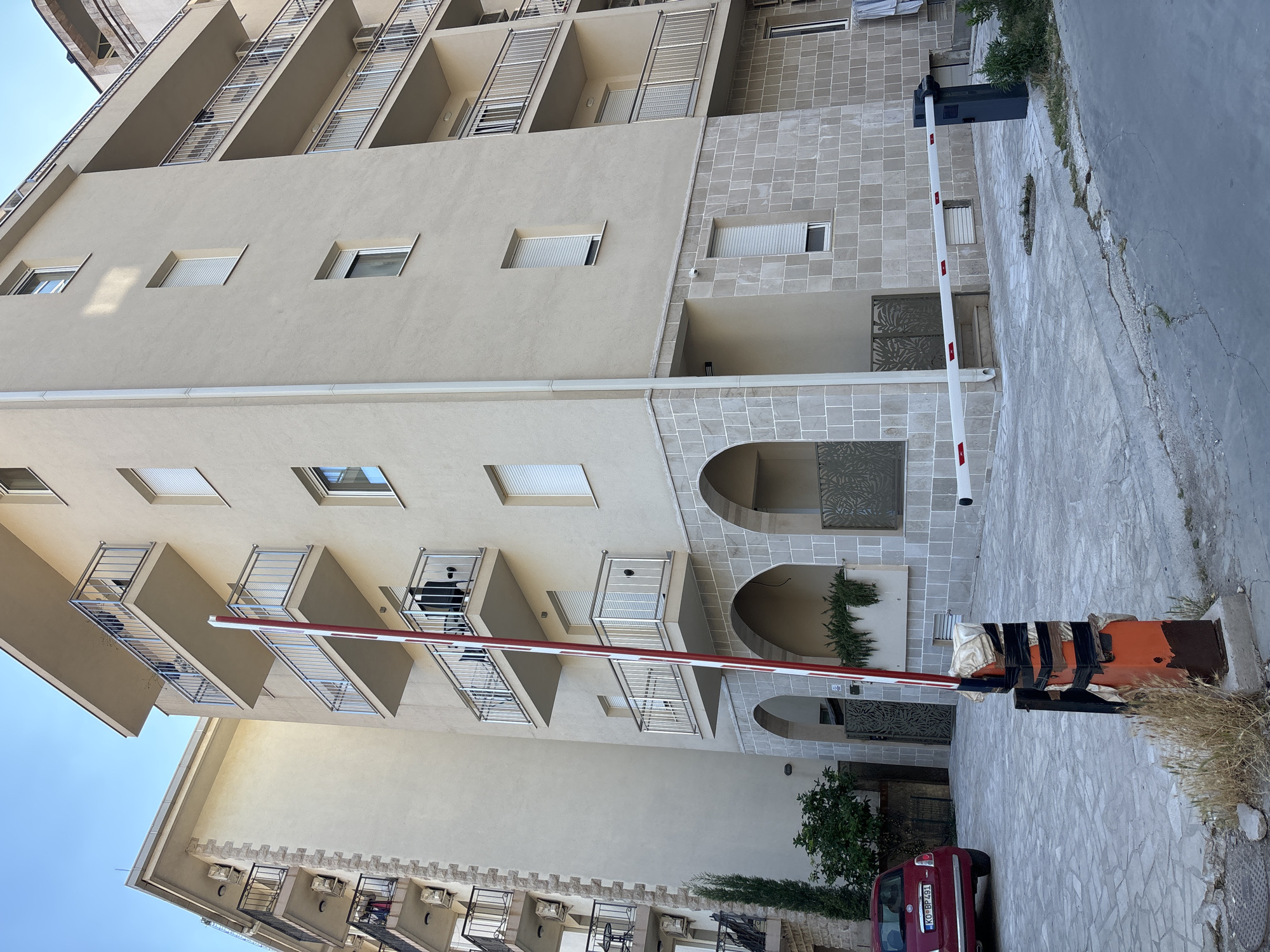 The image depicts a view of a multi-story apartment building with balconies, located in Budva, Montenegro. A red Fiat 500 car is parked in the foreground to the left. In the center, a retractable barrier gate is extended across a paved driveway, with its pole partially obscuring the building. The building features a light-colored facade and stone accents at the lower levels, with arched entrances. Balconies with metal railings are visible on multiple floors. The sky is clear and blue. The scene appears to be a residential area, possibly within a gated community or private parking lot.
