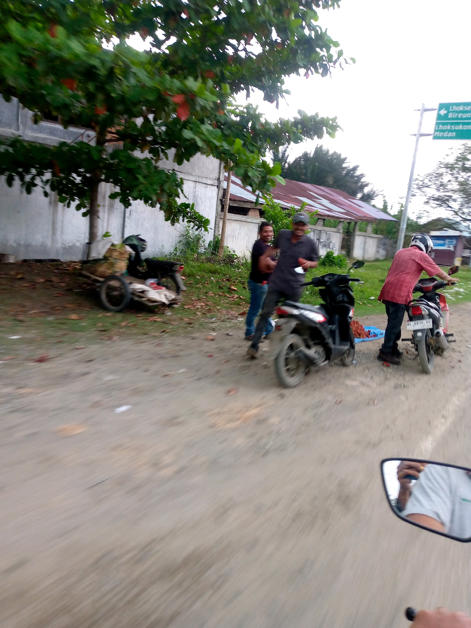 In Peurupok, Indonesia, on a dirt road, three individuals are present. A man in a red plaid shirt and helmet stands by a motorcycle. Another man in a black shirt and jeans is seated on a motorcycle. A third man, also in a black shirt, is standing next to him, holding a bottle. In the background, a cart filled with goods is attached to a motorcycle. A signpost points to Lhokseumawe, Bireuen, and Medan. A building with a reddish roof is visible behind the people. The scene is captured from a moving perspective, possibly from another motorcycle, as indicated by the reflection in a mirror. The environment appears to be a rural or semi-urban area.