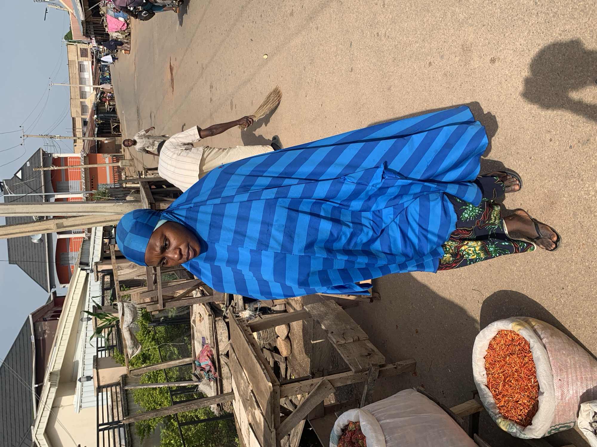 A medium shot shows a woman in a blue and black striped headscarf and a matching blue abaya walking on a street, likely in the city of Jos, Nigeria. She is looking towards her left with a focused expression. In the foreground to her left, a large sack filled with red chili peppers is visible. Behind her, a man is sweeping the dusty street with a broom, and further down the street, several other people are walking, some carrying goods. Buildings with corrugated iron roofs line the street, suggesting a market or commercial area. The overall scene depicts a bustling outdoor market environment under a clear sky.