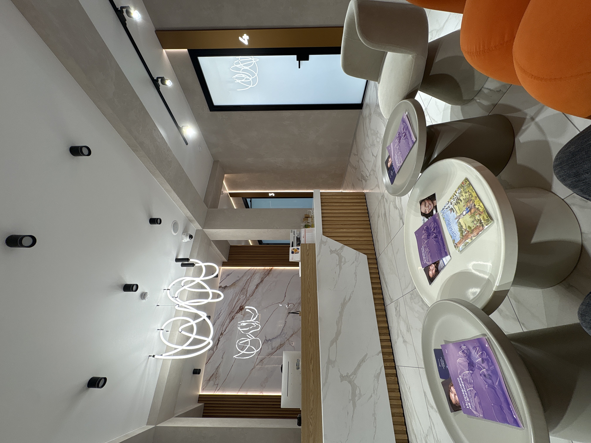 The image shows the waiting area of a clinic or office. The location may be in the city of Khabarovsk, Russia, as evidenced by the Russian-language magazine on one of the tables. The area features modern white and gray decor with marble accents. There are several round white tables with magazines on them, surrounded by white and orange chairs. The lighting includes recessed ceiling lights, track lighting, and decorative neon light fixtures on the wall. The floor is tiled, and there's a reception desk visible in the background. No people are clearly visible, although there are some legs and a foot.