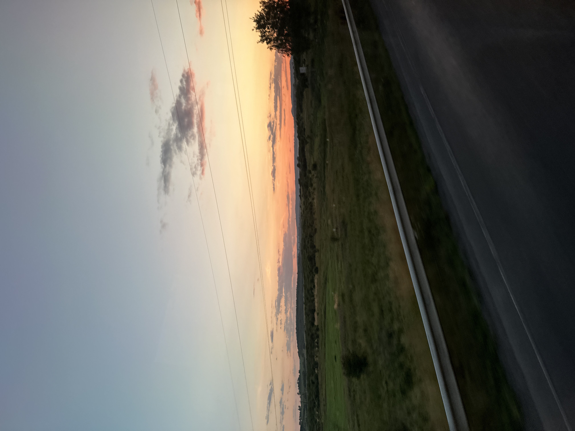 The image captures a pastoral landscape at dusk, likely near Chyzhykiv, Ukraine. The sky transitions from a pale blue at the top to a vibrant orange and pink sunset near the horizon, with scattered clouds adding texture. A dark silhouette of trees and rolling hills forms the backdrop. In the foreground, a lush green field stretches out, dotted with smaller trees and bushes. A silver guardrail runs along the bottom right of the frame, separating the field from a dark asphalt road, suggesting the vantage point is from a moving vehicle. Power lines are visible stretching across the sky. No people or specific buildings are discernible.