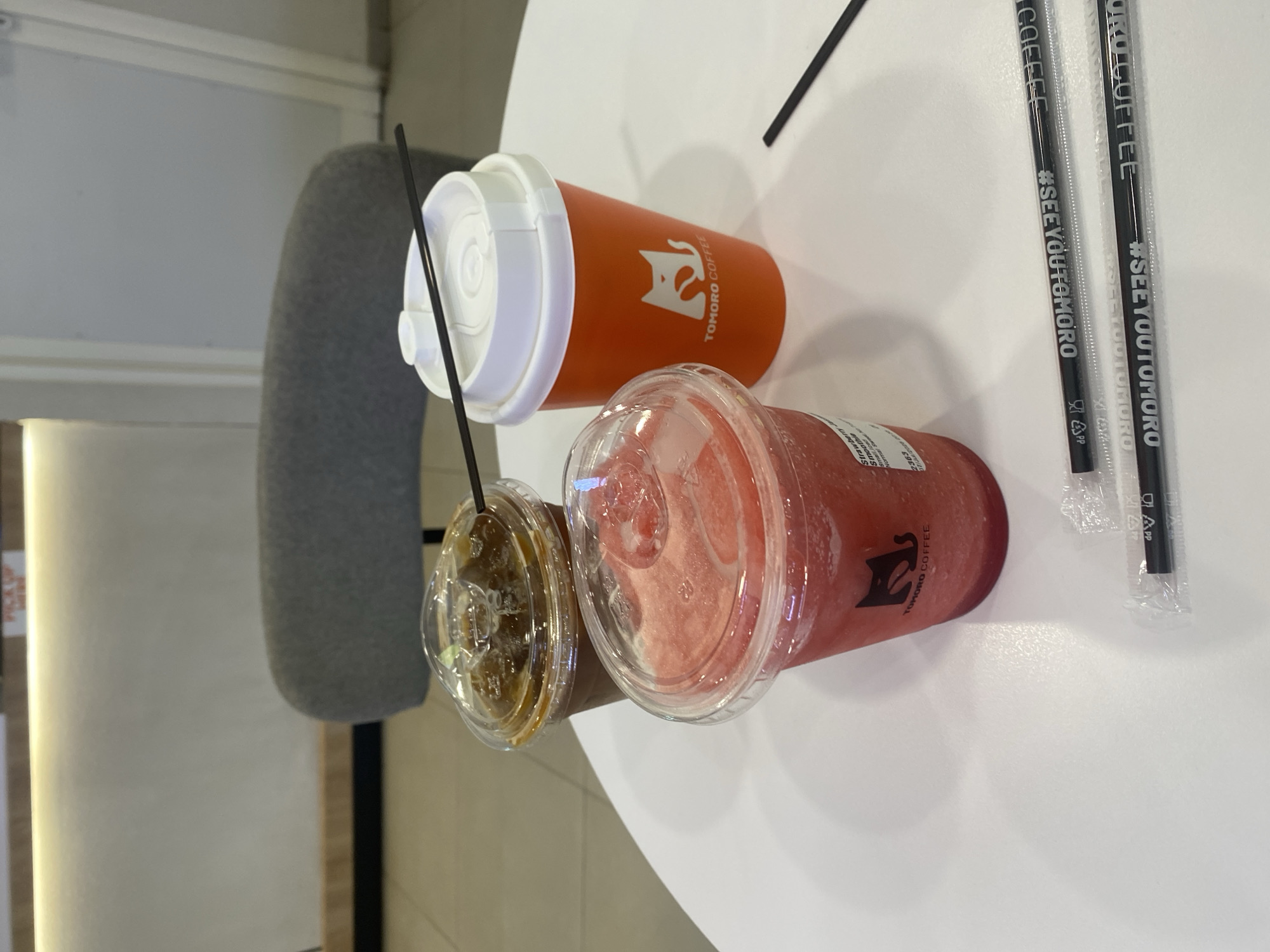 On a white, circular table, three beverages are arranged. The beverage in the foreground is a red, blended drink in a clear plastic cup with a domed lid, and appears to be a strawberry smoothie. The cup has black text and a black logo that reads "TOMORO COFFEE". To the right of the smoothie is an orange paper cup with a white lid and a black straw. The cup also features the "TOMORO COFFEE" logo. In the background, to the left, is a clear plastic cup filled with a dark brown beverage, presumably coffee, with ice and a straw. Two black plastic straws with white text are lying on the table in front of the beverages. The table is set in front of a grey upholstered chair. In the background, a light-colored counter with a yellow accent light is visible, along with a portion of a wall and a door. The scene is set in Banjarbaru, Indonesia.