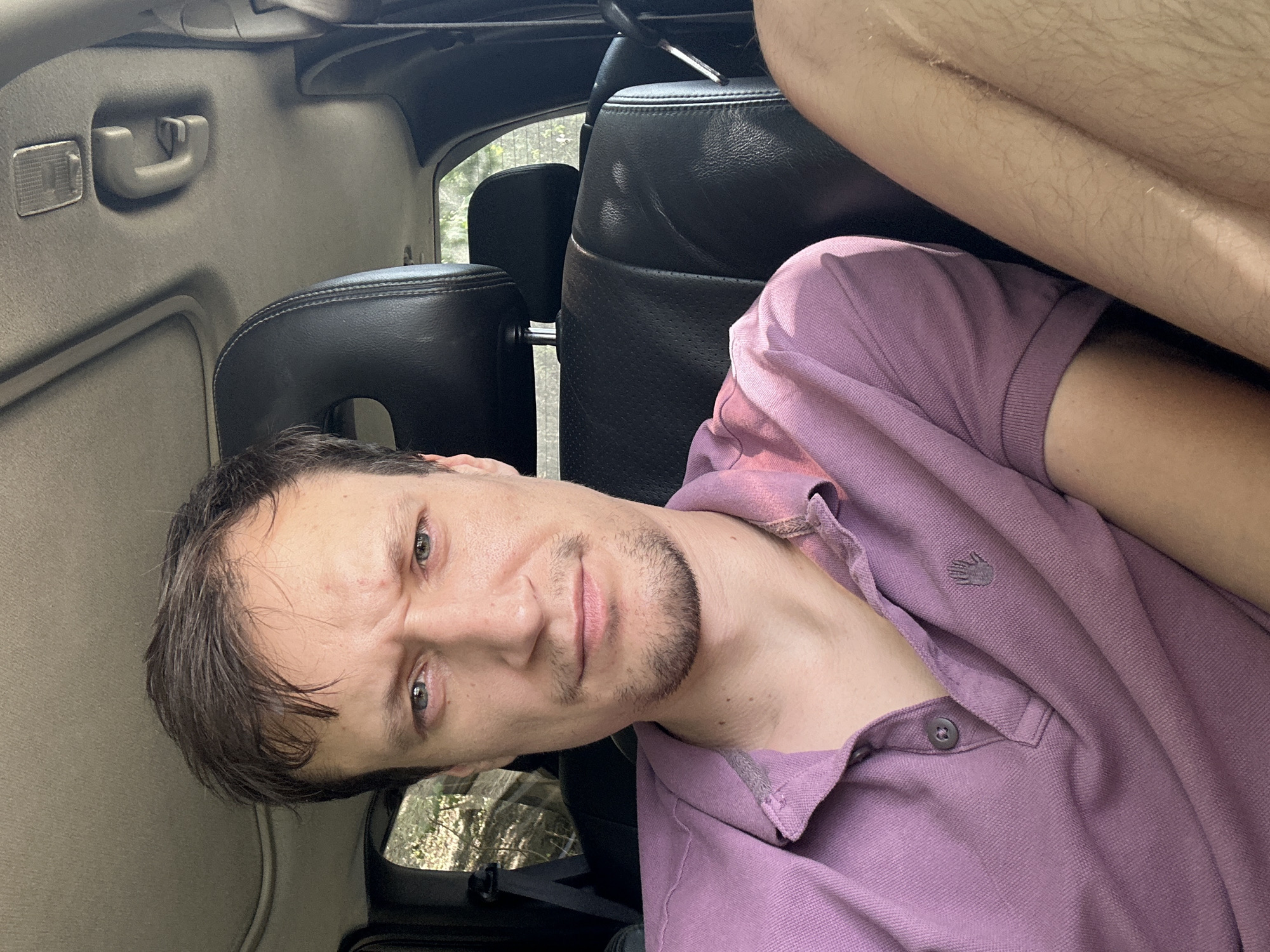 A man is sitting in the passenger seat of a car, looking directly at the camera. He has short, dark hair and is wearing a purple polo shirt. The interior of the car is visible, including the black leather seats and the car's ceiling. The setting appears to be outdoors, with greenery visible through the car window. The man's expression is stern, with his eyebrows furrowed. The location of Makiv, Ukraine, is not discernible from the image itself.