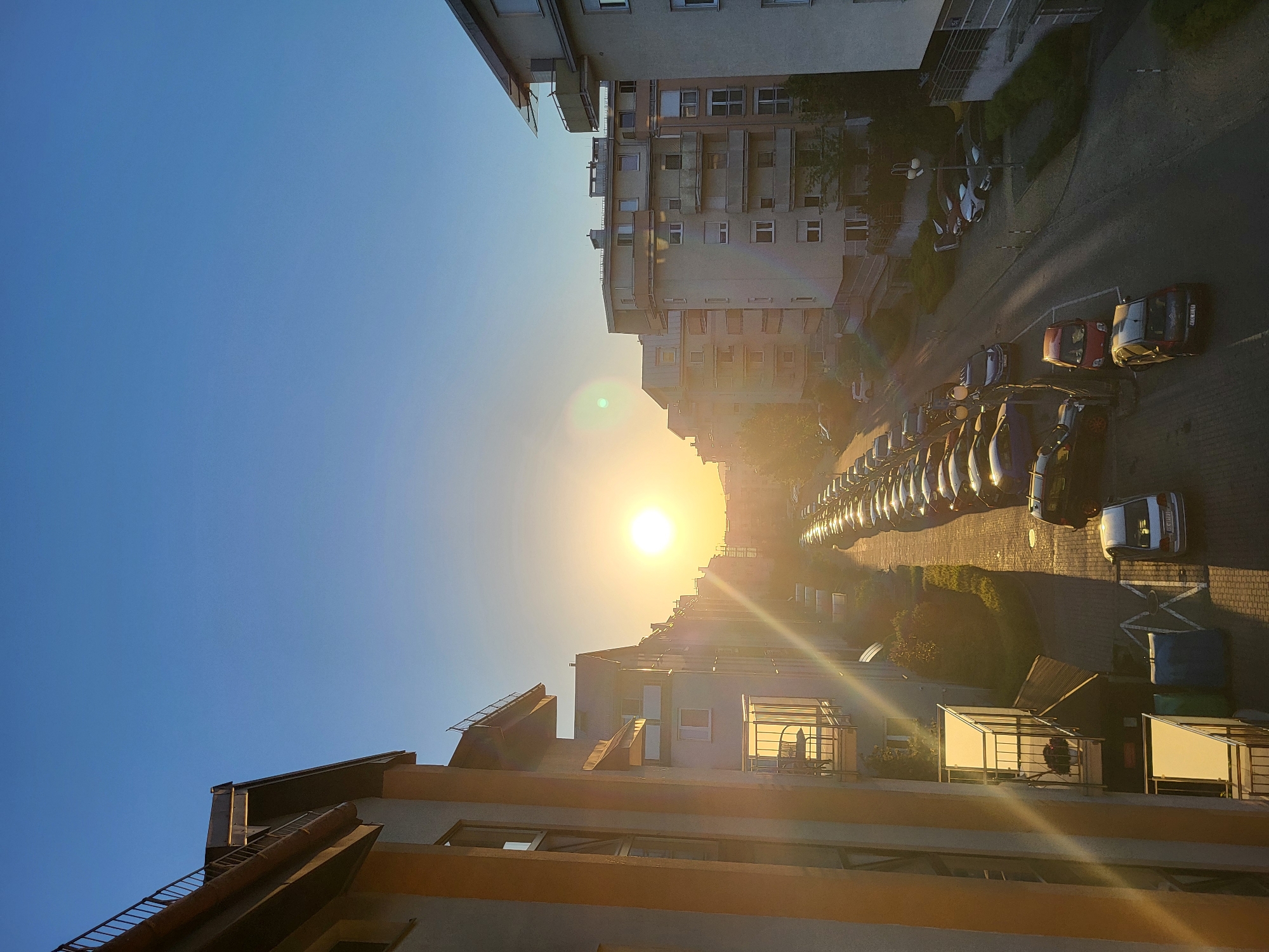 The image presents an upward view in a cityscape. The sun is positioned centrally, appearing low on the horizon. Rows of parked cars line a street flanked by buildings of varying heights. The buildings are constructed in concrete and appear residential or commercial. The sky is predominantly clear and blue.