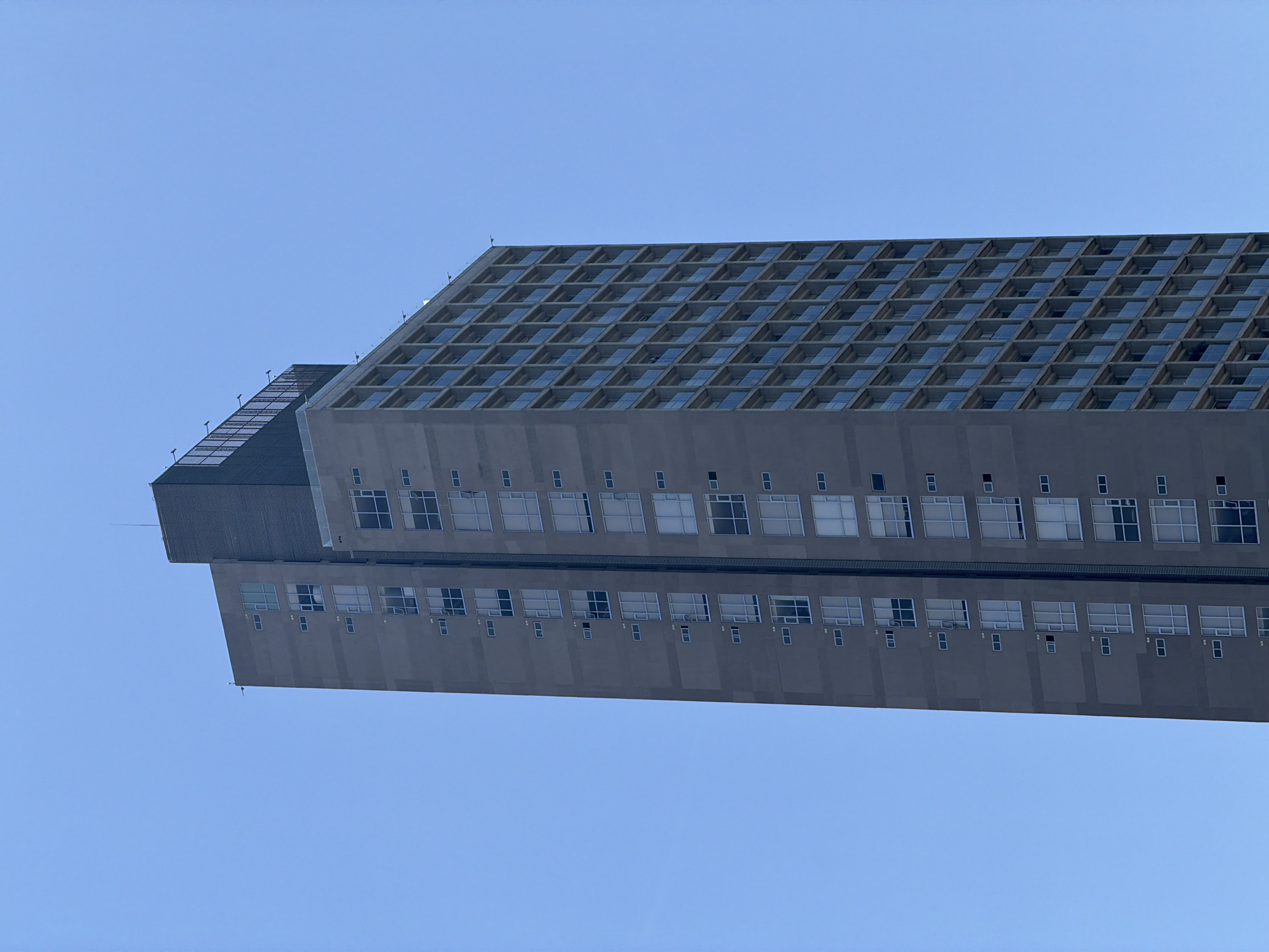 The image captures a modern architectural structure against a clear, vibrant blue sky. The photograph is oriented vertically, presenting the building horizontally across the frame, as if it were a high-rise viewed from directly below and then rotated. When conceptually re-oriented, it depicts a building seen from a low-angle perspective, looking upwards.
The prominent subject is a large, predominantly gray building. Its uppermost section features a distinctive, large-scale grid pattern, where each square or rectangular recess appears to contain a pane of glass, suggesting a skylight system or a unique facade design. To the left, a smaller, darker section of the building juts out, also exhibiting a grid pattern, possibly composed of solar panels or a similar material.
The main body of the building below this top section displays multiple rows of windows. These windows vary in size, with some appearing as small square openings and others as larger, rectangular panes, creating a structured but varied appearance along the building's side. The overall construction material appears to be concrete or a similar smooth, monochromatic substance.
The sky is uniformly bright and clear, indicating fair weather and daytime conditions. There are no clouds visible, emphasizing the stark lines of the architecture against the open expanse. No people, discernible activities, or visible text are present in the image. The scene is located in Puebla, Mexico.