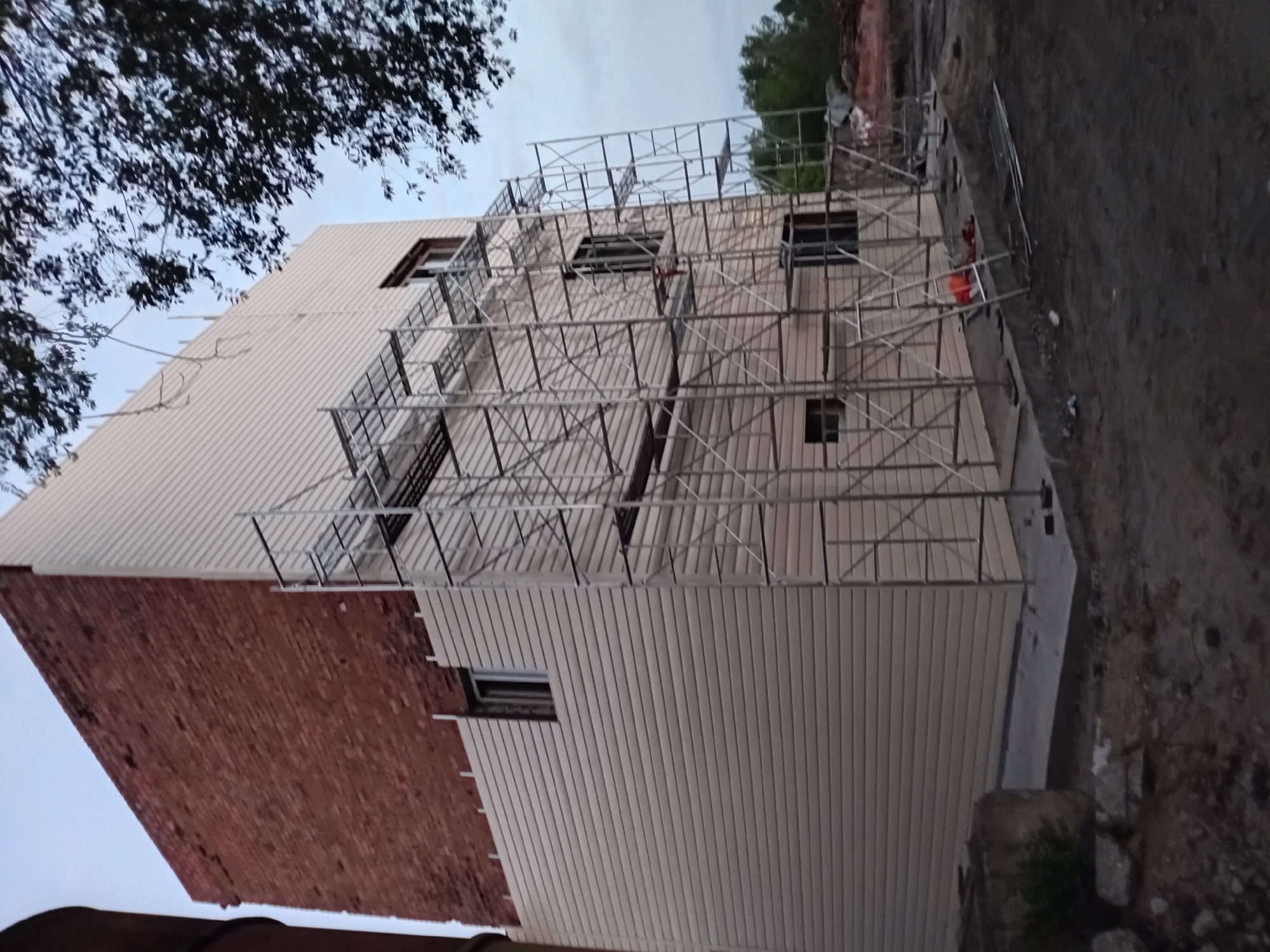 The image depicts a multi-story building under construction in the city of Semey, Kazakhstan. The building has a brick upper section and a beige, horizontally-paneled lower section. Extensive scaffolding is erected around the lower portion of the building, indicating ongoing construction or renovation work. Several windows are visible on both the brick and paneled sections. The ground surrounding the building appears to be a construction site with loose soil and debris. Trees are visible in the background, and the sky is overcast, suggesting it is either early morning or late evening. No people are visible in the image.