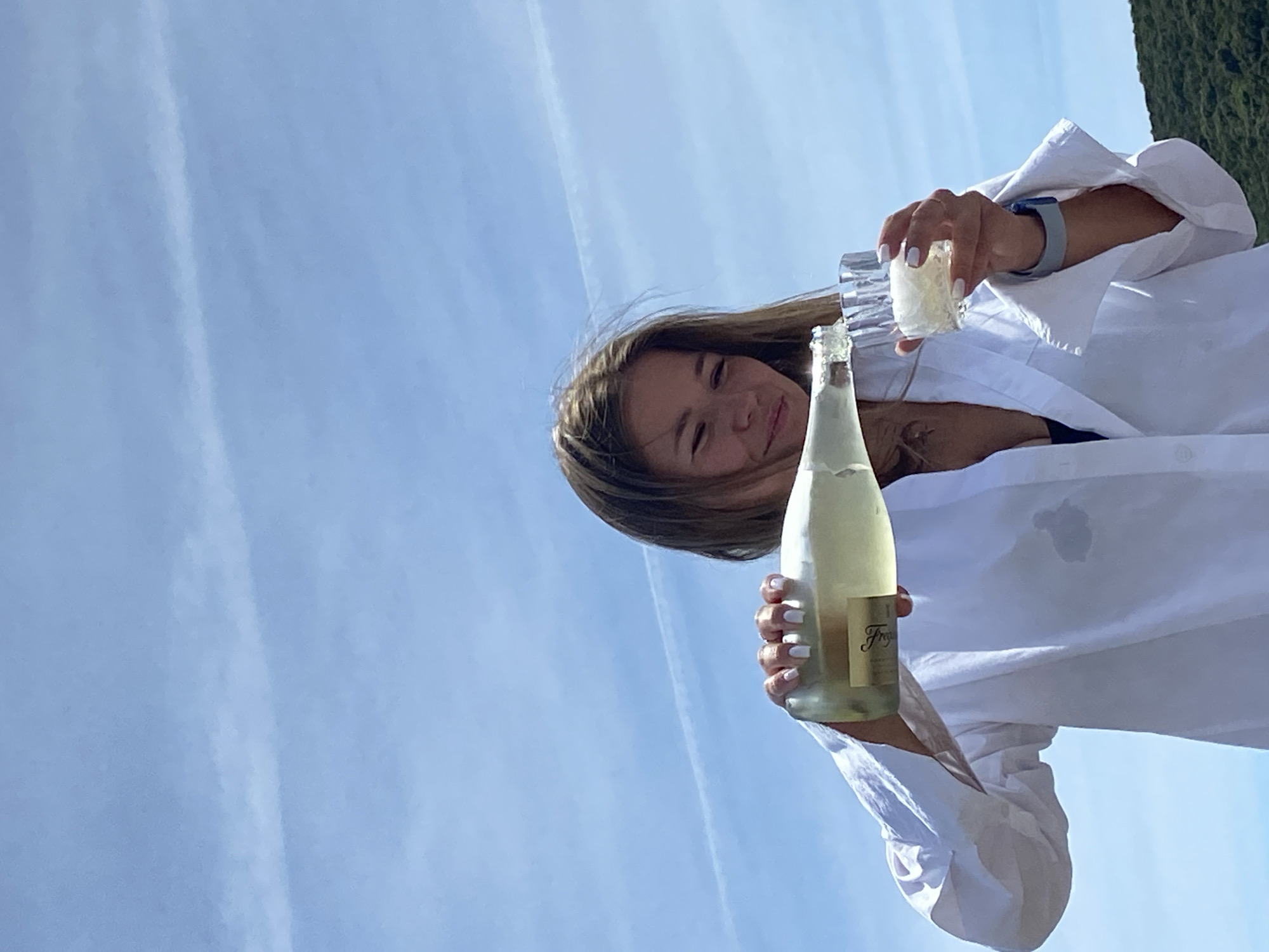 A young woman stands outdoors on a sunny day, pouring sparkling wine from a Freixenet bottle into a glass. She wears a white button-down shirt and a watch. The backdrop is a clear blue sky with faint contrails and some greenery on the right side of the image. The location is Prijevor, Montenegro.