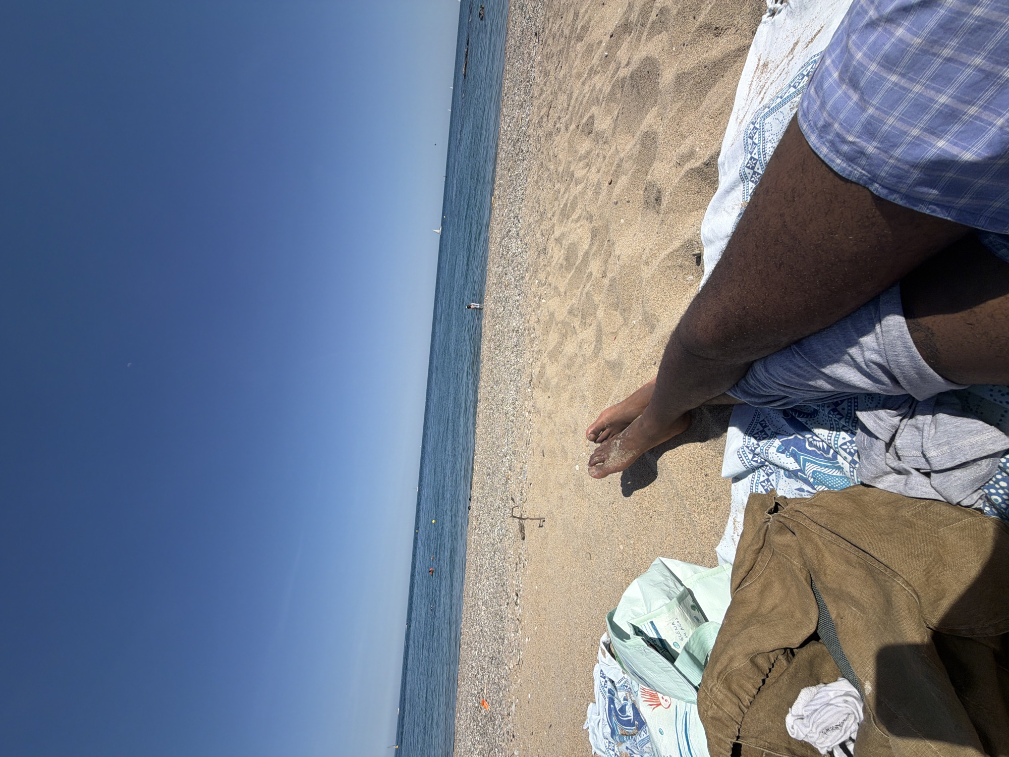 The image shows a person relaxing on a beach in Sant Adrià de Besòs, Spain, next to the Mediterranean Sea. The sky is clear and blue. The beach consists of sand and pebbles. The person is wearing a blue plaid shirt and grey shorts. A brown jacket and a light green shopping bag are lying on a blanket next to them. The person's feet are visible, resting on the sand. Several sailboats can be seen on the water in the distance.