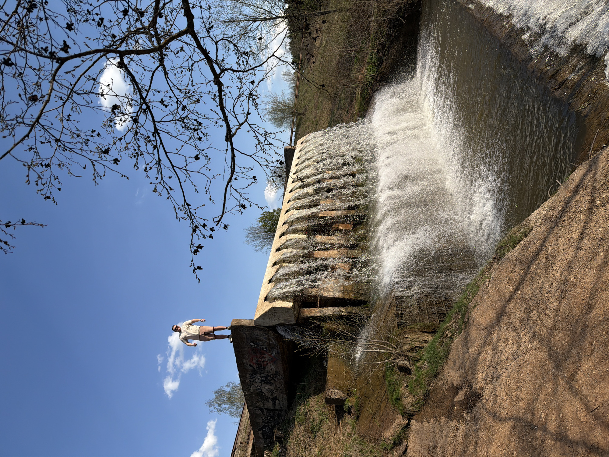 A man stands on a concrete structure adjacent to a cascading waterfall. The scene includes a clear blue sky with scattered clouds and tree branches in the upper left. The waterfall appears to be man-made, with water flowing over a stepped concrete barrier. Vegetation covers the banks surrounding the waterfall. The image's perspective is tilted, possibly captured in Kamennoye, Russia.
