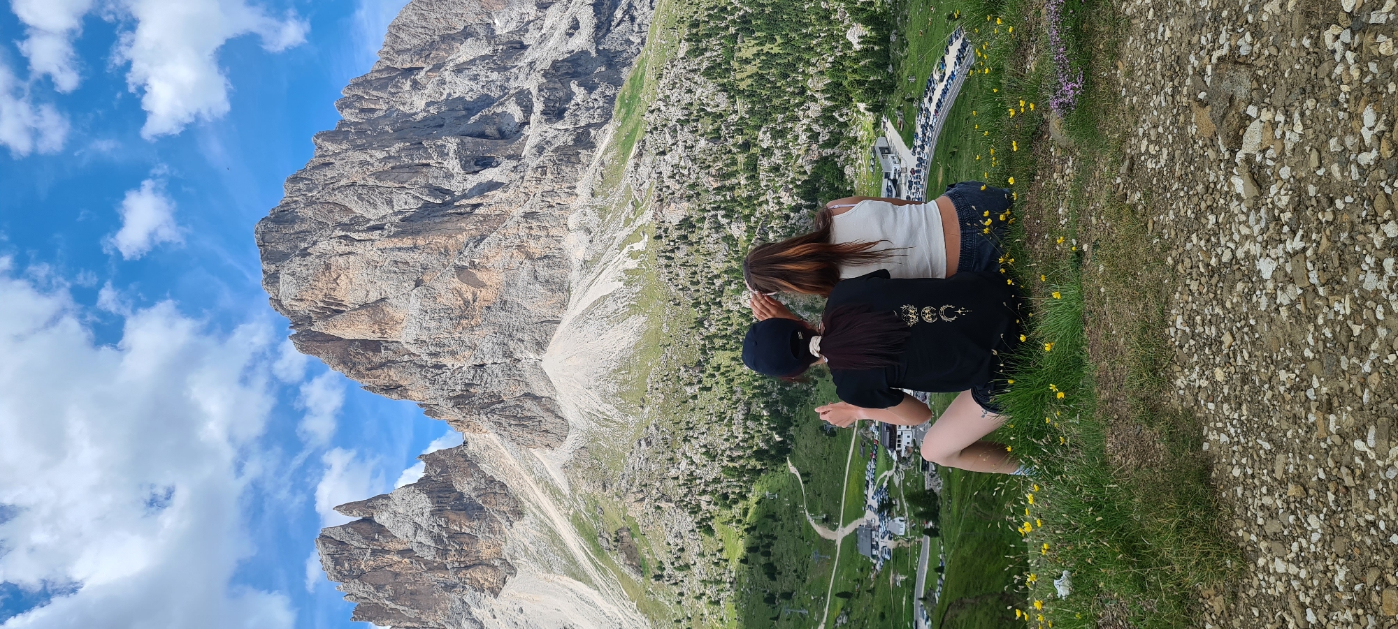 Two women sit facing away from the camera, looking out at a vast mountainous landscape. The woman on the left wears a black t-shirt with a graphic design and a black baseball cap. The woman on the right wears a white tank top. They are seated on a grassy hill dotted with yellow wildflowers. Below them, a winding road leads to a parking lot filled with cars, and several buildings. The background is dominated by massive, rugged mountains with steep, rocky slopes, partially covered in green vegetation and trees. The sky is blue with scattered white clouds. The location is in Val Gardena, South Tyrol, Italy, likely near the Sella Pass, given the distinctive mountain formations.