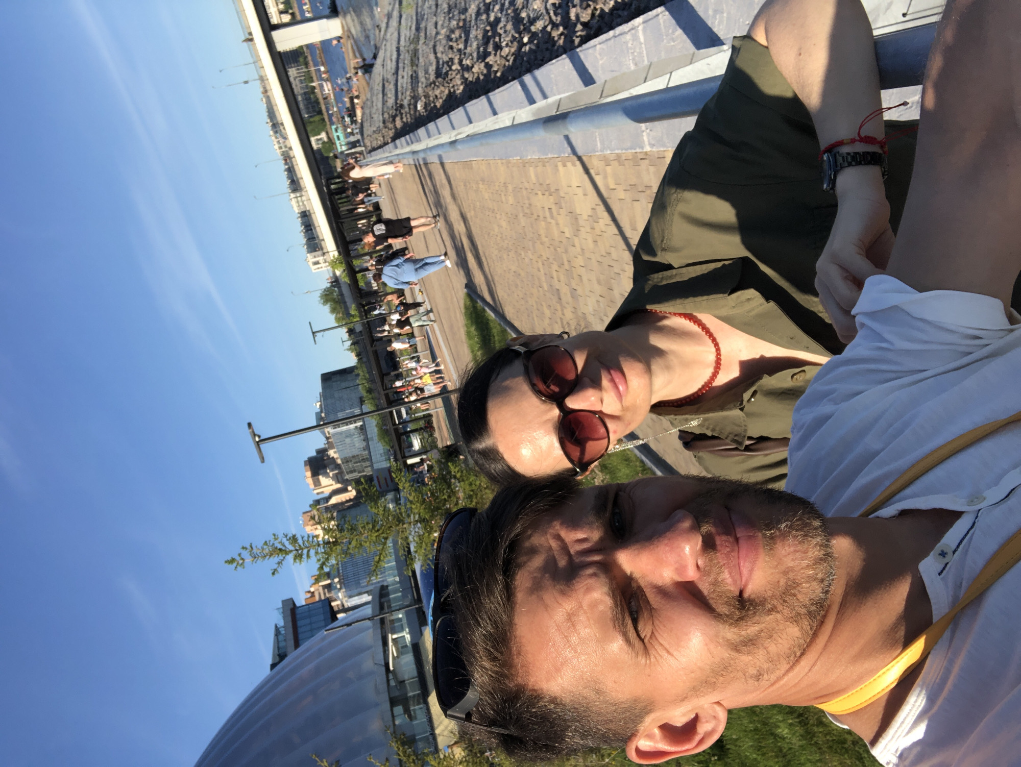 In Saint Petersburg, Russia, on a sunny day, a man and a woman take a selfie with a river and urban landscape in the background. The man, in a white shirt, is in the foreground, with sunglasses perched on his head. The woman, wearing a green shirt and sunglasses, is beside him, leaning on a railing. People are visible in the background, walking along a paved path near the water. A modern building with a curved roof is to the left, and a bridge spans the river in the distance. The scene appears to be a waterfront promenade.