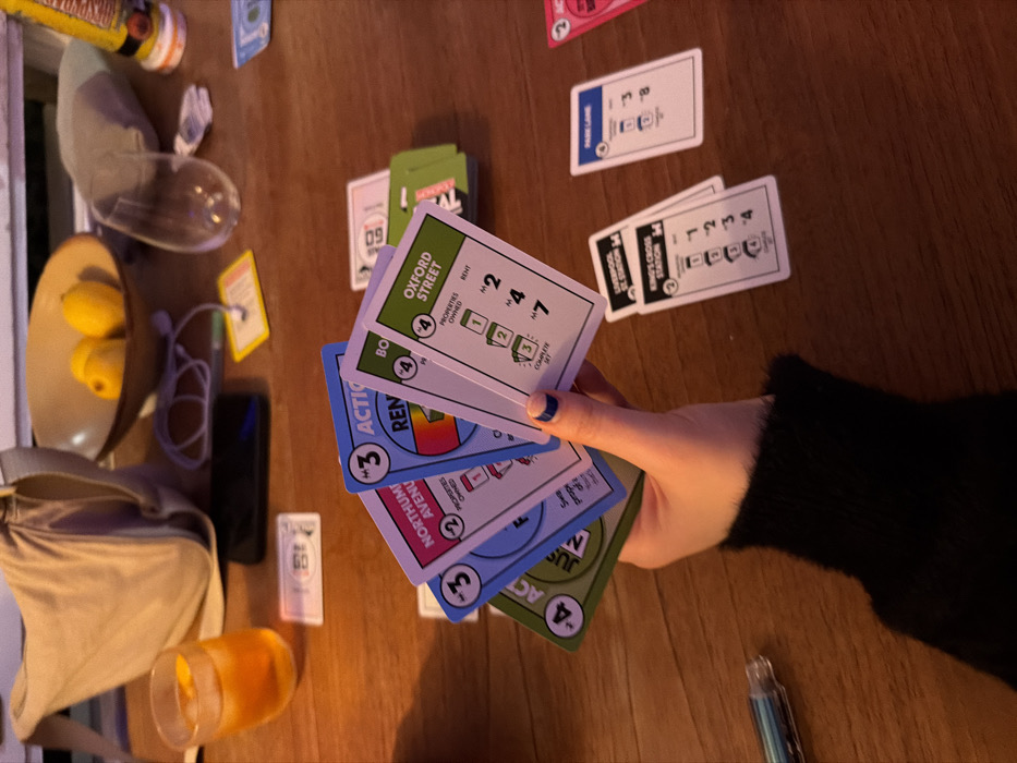 The image presents a close-up, overhead perspective of a polished wooden table under warm, ambient lighting, focusing on a card game. A person's left hand, with a dark blue-tipped index fingernail and a dark, fuzzy sleeve cuff, holds a fan of five Monopoly Deal cards. Visible cards in hand include a green "JUST SAY NO!" card (value 4), a blue "NORTHUMBERLAND AVENUE" property card (value 3), a blue "ACTION RENT" card (value 3), and a green "OXFORD STREET" property card (value 4).
On the table surface, various other Monopoly Deal cards are arranged: a stack of green property cards, a pink property card (partially visible), a blue "PARK LANE" property card (value 4), and two stacked railway station cards identifying "LIVERPOOL ST. STATION" and "KING'S CROSS STATION" (both value 4). A single yellow "M1" house card and a "M5" bill card are also visible.
Ancillary objects on the table include a clear glass containing an amber beverage with ice, a black smartphone lying face down, a light-colored fabric sling bag, a beige bowl holding two yellow lemons, an inverted empty clear wine glass, a small grey fabric pouch, and a clear-bodied pen with blue internal components. The presence of "NORTHUMBERLAND AVENUE", "OXFORD STREET", "PARK LANE", "LIVERPOOL ST. STATION", and "KING'S CROSS STATION" on the game cards identifies specific locations within London, United Kingdom.