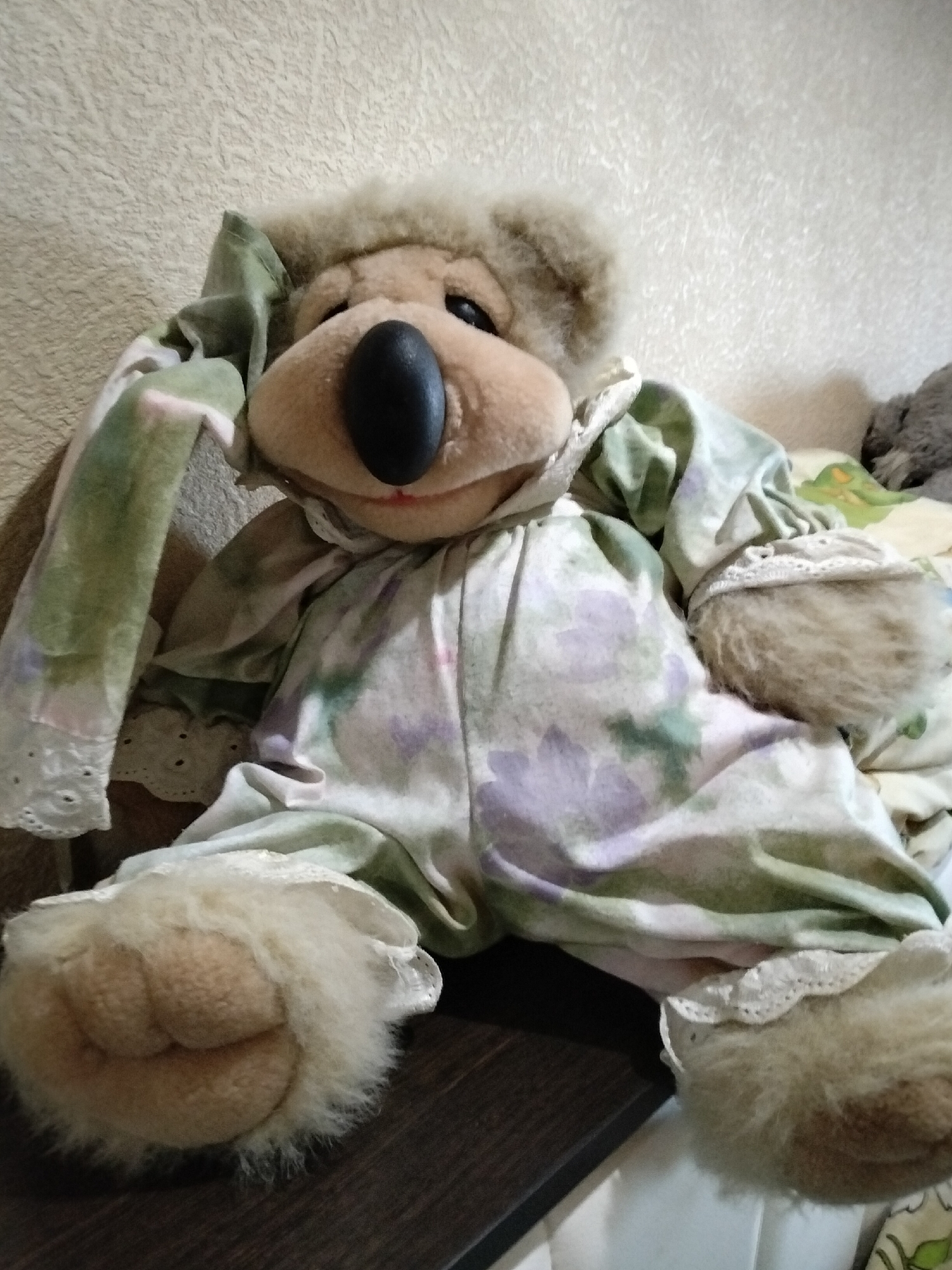 A light brown teddy bear with a large black nose is sitting on a dark brown surface. The teddy bear is wearing a light green and purple floral patterned pajama outfit with lace trim. The background is a textured beige wall. The image is taken indoors, likely in a home environment, and there is no indication of a specific city or location.