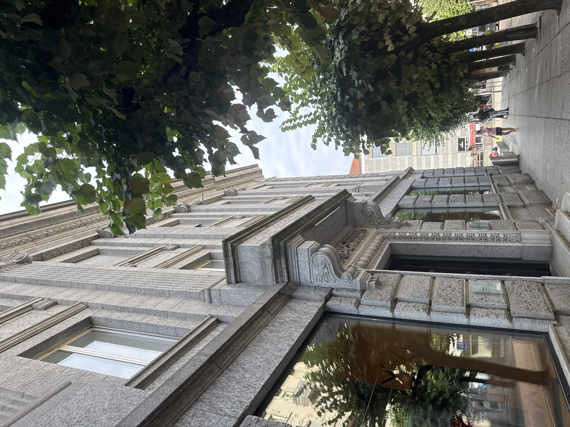 The image captures a street scene in Bellinzona, Switzerland, featuring a grand, ornate building constructed of granite blocks, likely a historical structure or bank given the inscription "BPS SUISSE" above an entrance. Trees with lush green foliage line the sidewalk in front of the building, casting shadows. A sidewalk runs parallel to the building, with a few individuals walking. On the right side of the image, a woman in a pink t-shirt and shorts is walking towards the viewer, while a man in a dark jacket and jeans walks away. In the background, other buildings and a street with parked cars are visible. The sky is overcast.