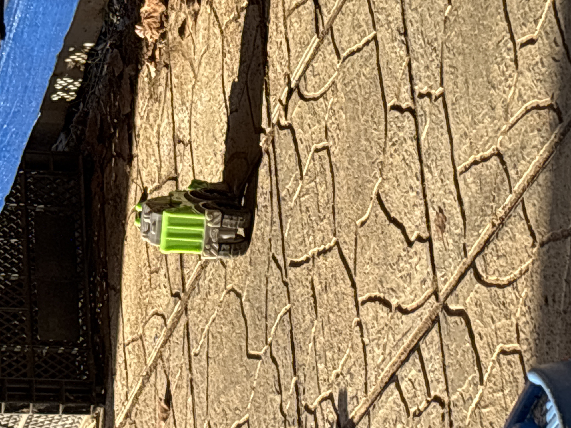 A green and gray toy vehicle lies on a patterned stone surface. The surface appears to be a walkway or patio area, judging by the repeating pattern of the stones. A portion of a blue tarp or sheet is visible in the top left corner, partially covering a black lattice object. Dried leaves are scattered on the ground to the left of the toy. There are no people visible. The location is not identifiable as Chaikovsky, Russia based on the given image.