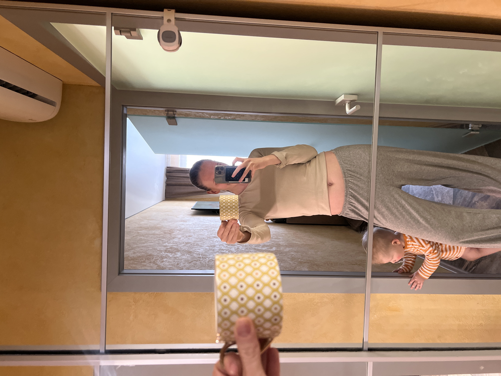 The image shows a mirrored scene reflecting a man taking a selfie with his phone while holding a yellow and white patterned cup. He's wearing a beige long-sleeved shirt and gray sweatpants. A baby in an orange and white striped shirt is crawling near the bottom of the reflection. The room behind them appears to be a living space with a neutral color palette. The location is likely indoors, specifically in a bathroom or closet area due to the mirrored sliding doors. There is no clear information that suggests the image was taken in Khimki, Russia, therefore that detail cannot be included in the response.