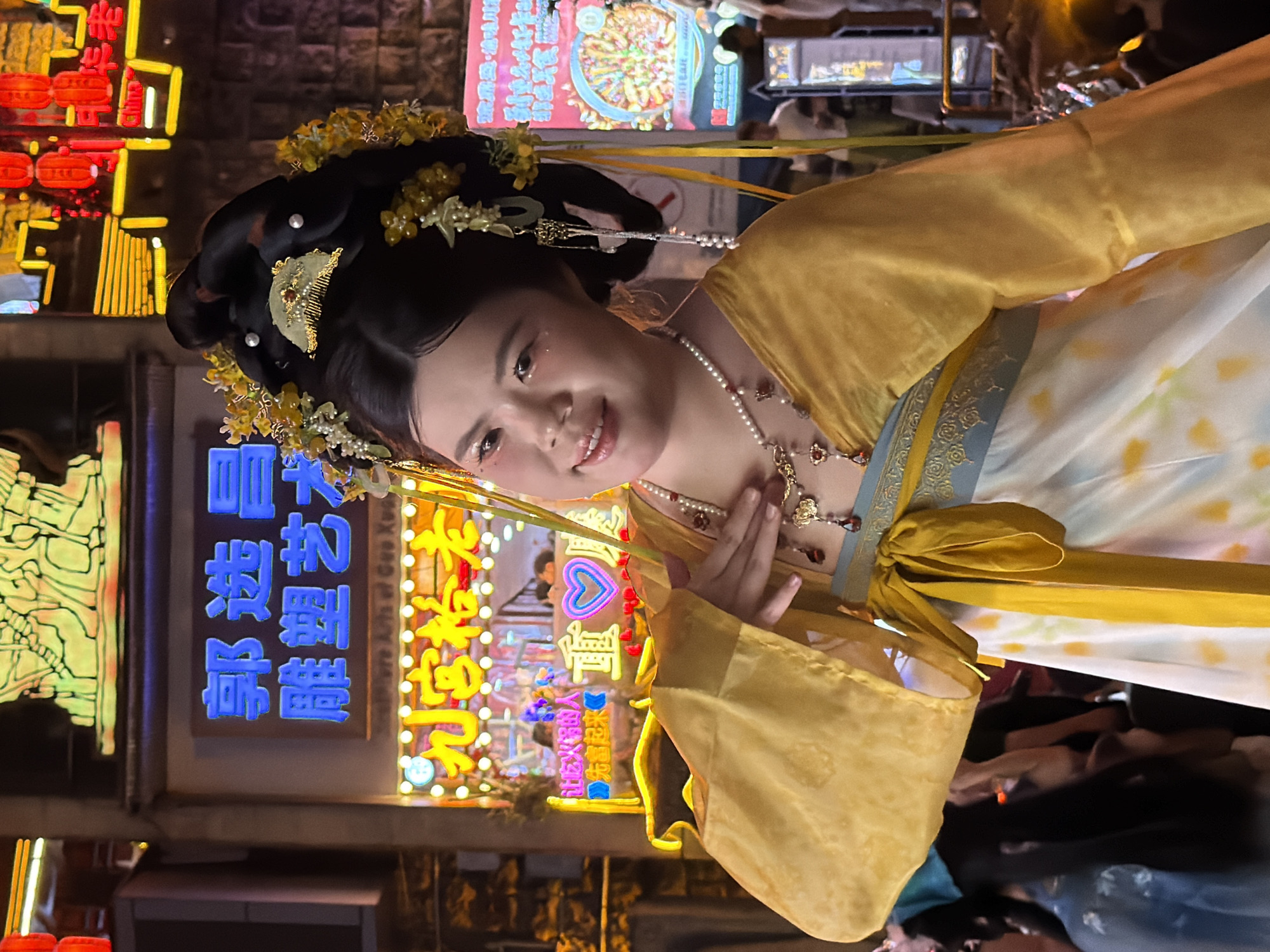 A young woman in traditional Chinese attire with elaborate hair ornaments and makeup poses in front of a brightly lit neon sign. The sign, in blue and yellow Chinese characters, appears to be for a business related to arts and crafts or food. The woman is wearing a yellow robe over a white dress with floral patterns, adorned with pearl necklaces and intricate jewelry. Her left hand is raised to her chest, and she is smiling at the camera. In the background, blurred figures and more neon signs indicate a bustling urban environment, likely a street in Yuzhong District, China, known for its vibrant nightlife and commercial activity. The overall scene suggests a festive or cultural event.