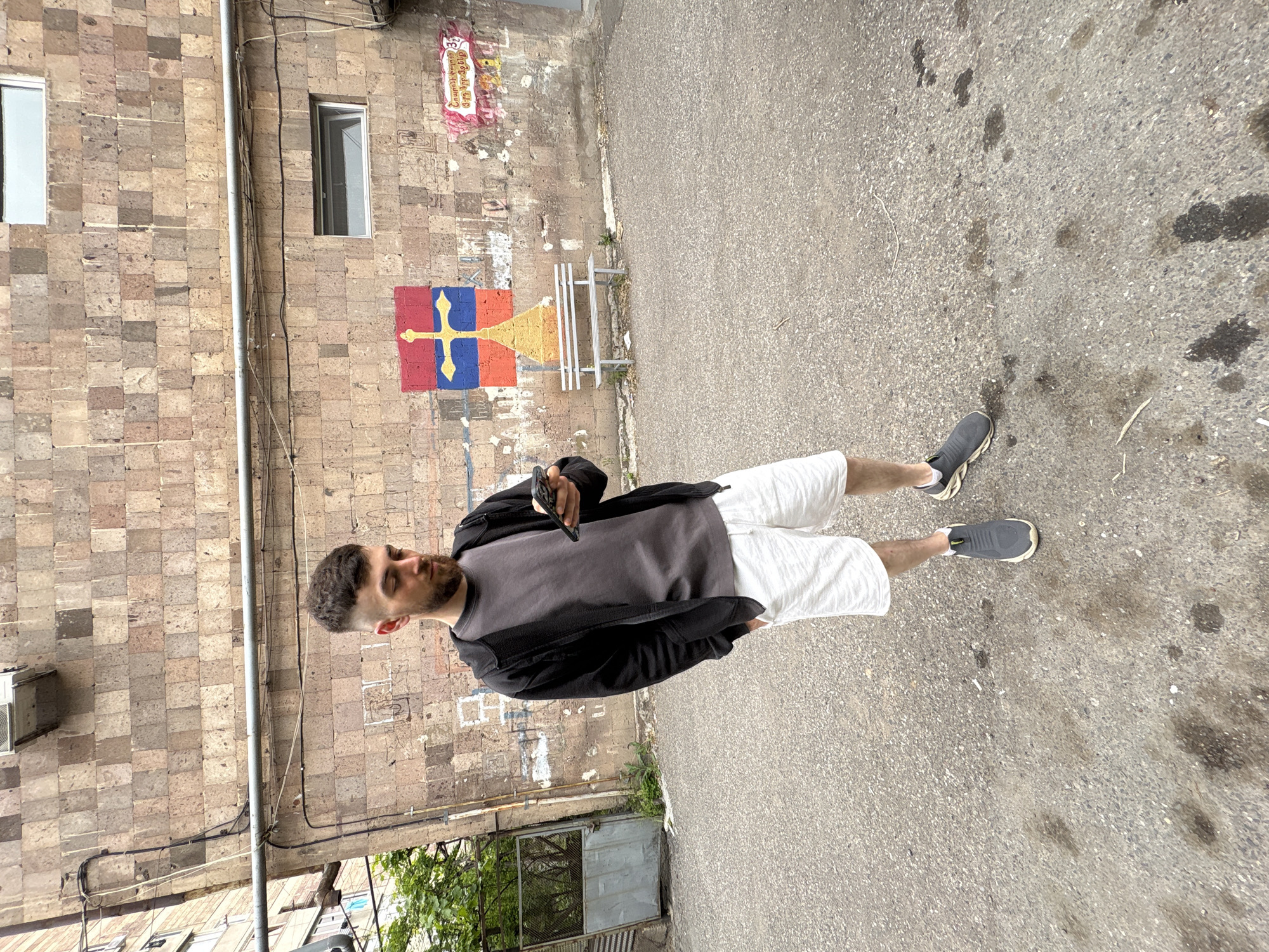 The image captures a man standing in an alleyway, likely in Yerevan, Armenia given the architectural style and painted motifs on the wall. The man is wearing a grey t-shirt, white shorts, a black jacket, and grey sneakers. He is holding a mobile phone. The background includes a brick building with two windows, electrical wiring, and a pipe running vertically along the wall. Painted on the brick wall is a motif featuring colors of red, blue, orange, and yellow, similar to the Armenian flag. A metal bench sits nearby. The ground is paved and shows some staining.