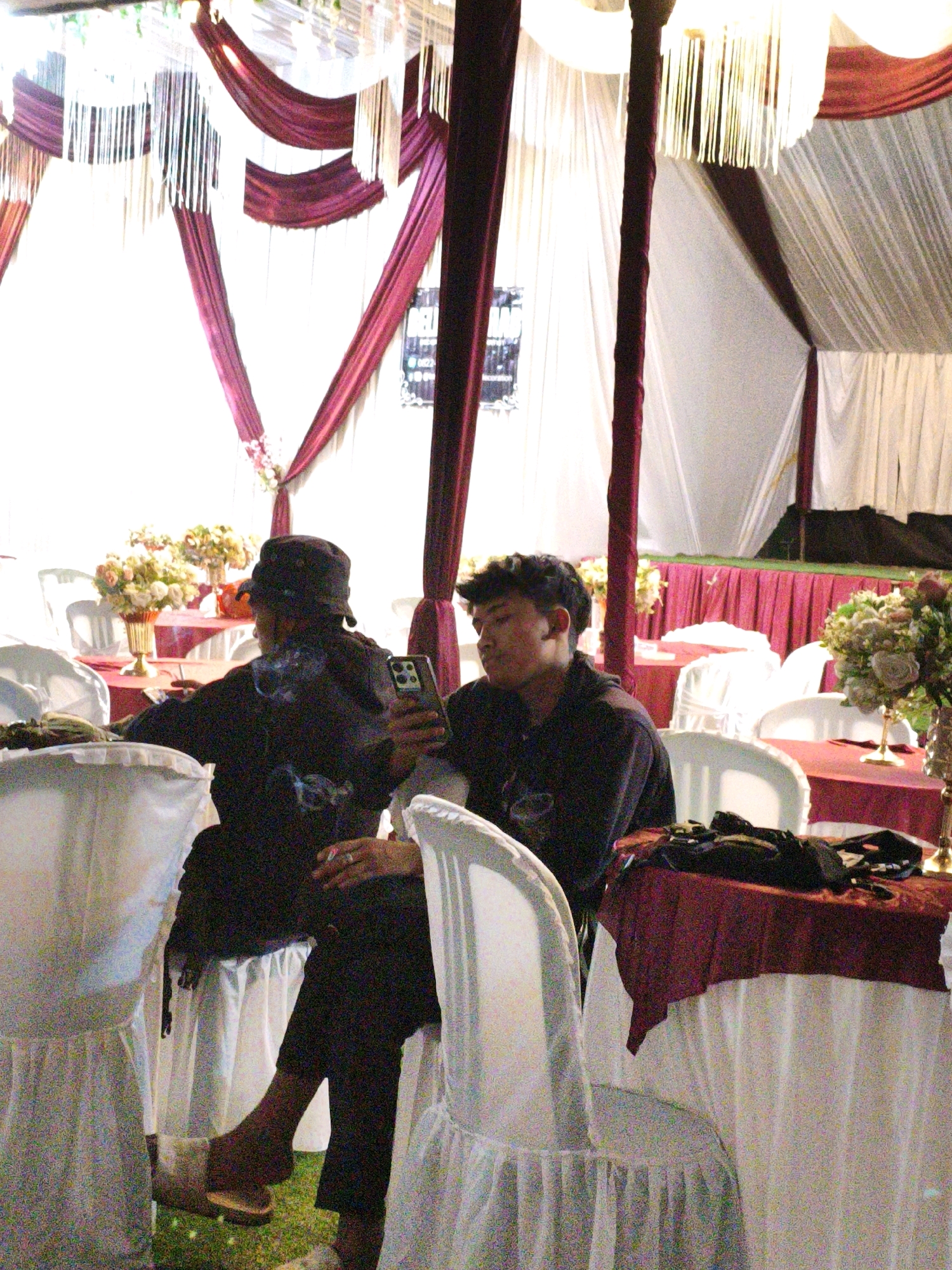 The image depicts an indoor event, likely a reception or gathering, decorated with draped burgundy and white fabric, as well as hanging fringe accents. Two individuals are seated at tables covered with burgundy tablecloths. One person, facing away from the camera and wearing a dark hat and clothing, is smoking and holding a cigarette. The second person, facing the camera and wearing a dark hooded jacket, is holding a smartphone. In the background, several round tables and white-covered chairs are visible, along with floral arrangements. The scene appears to be set up in a covered area, possibly a tent, with natural light filtering in from the sides. There are no specific visual cues to indicate the location is Lindung Jaya, Indonesia.