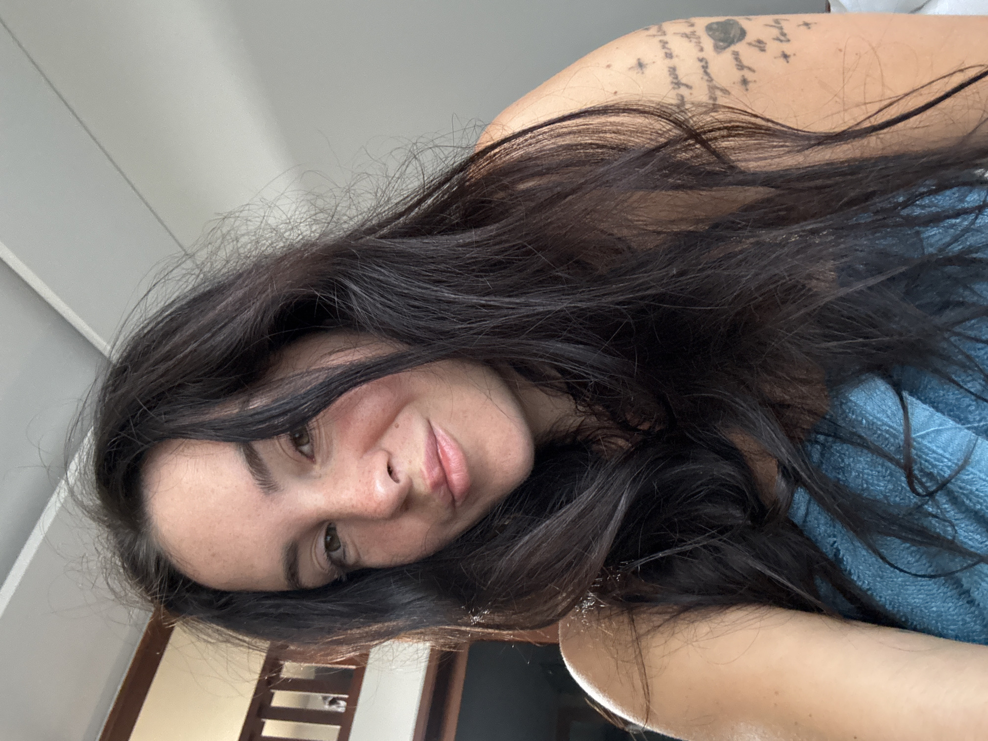 In the city of North Kuta, Indonesia, a young woman with long, dark, wavy hair is captured in a close-up, selfie-style portrait. She has fair skin with freckles visible across her nose and cheeks. Her eyes are a light brown, and she has a subtle smile on her lips. She appears to be wearing a blue towel or garment, draped around her shoulders. On her upper left arm, a tattoo is partially visible, featuring text and a graphic of a planet. The background is blurred, suggesting an indoor setting, with hints of a white ceiling and a wooden structure with railings. The lighting is soft, illuminating the woman's face from the front.