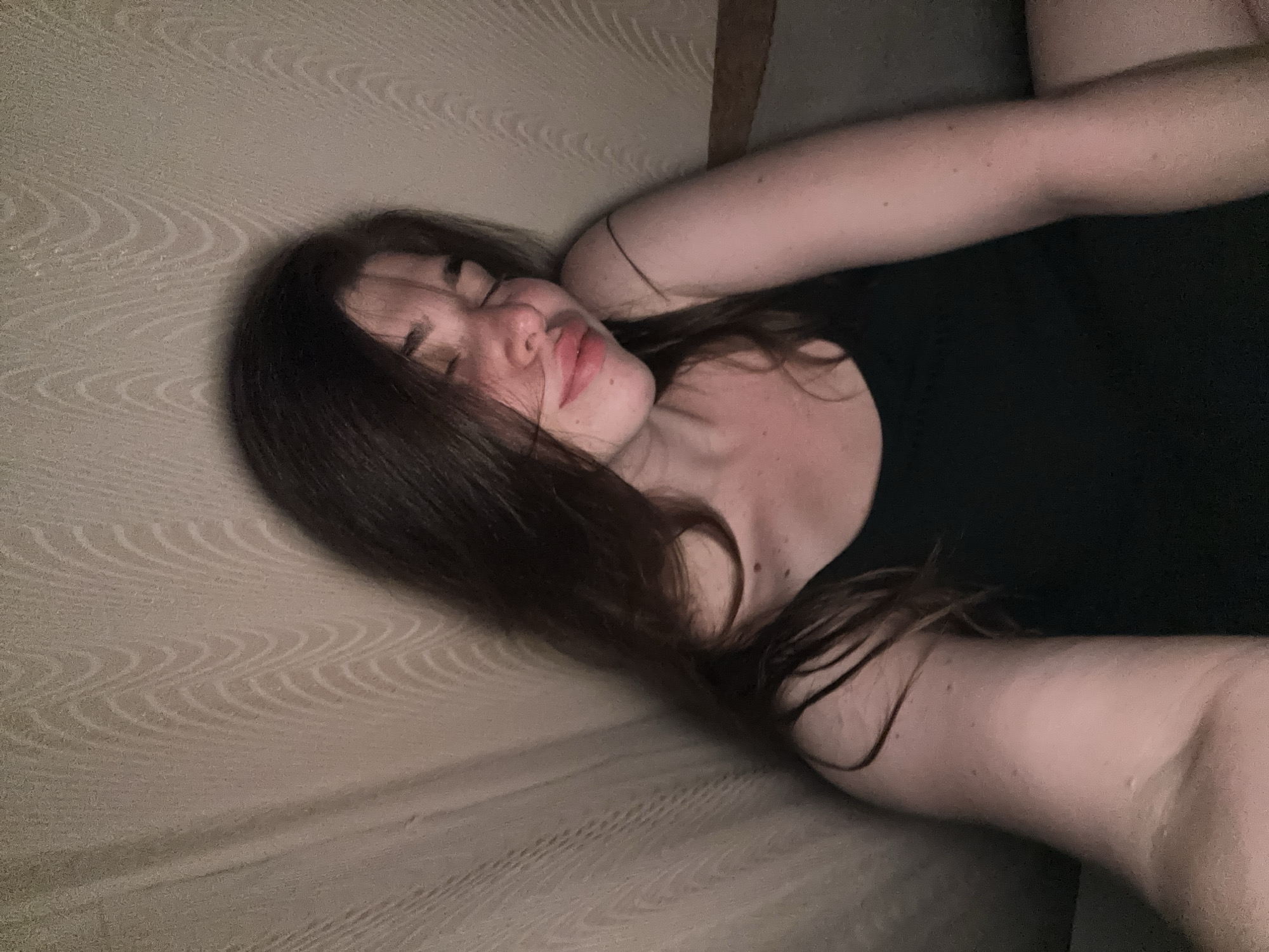 A young woman with long, dark hair is captured in a close-up, selfie-style shot. Her eyes are closed, and her head is tilted slightly, with her lips parted in a soft smile. She is wearing a dark, sleeveless top. The background appears to be a textured wall or fabric in a neutral tone, with soft lighting creating a gentle shadow on the left side of the frame. The overall mood is relaxed and intimate. The image is not geo-tagged and no information about the city of Мещёры, Russia is present in the visual data.