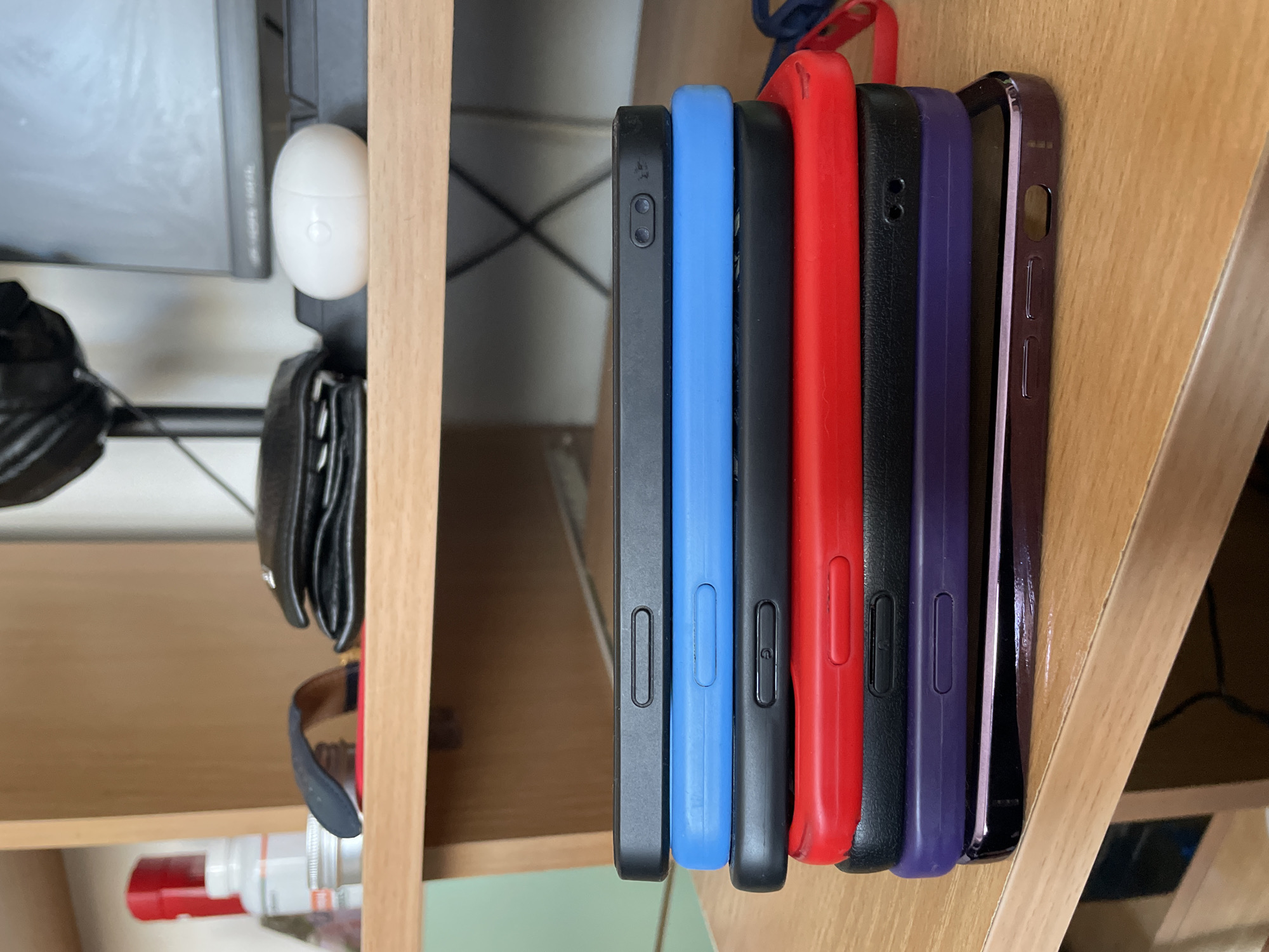 The image shows six mobile phones stacked vertically on a wooden surface. The phone cases are various colors: black, blue, black, red, black, purple. Behind them, a section of a shelf unit is visible, containing items like headphones, a wallet, sunglasses, and a bottle. The setting appears to be indoors, likely a home environment. There is no information to suggest that the location is in the city of Asenovgrad, Bulgaria.