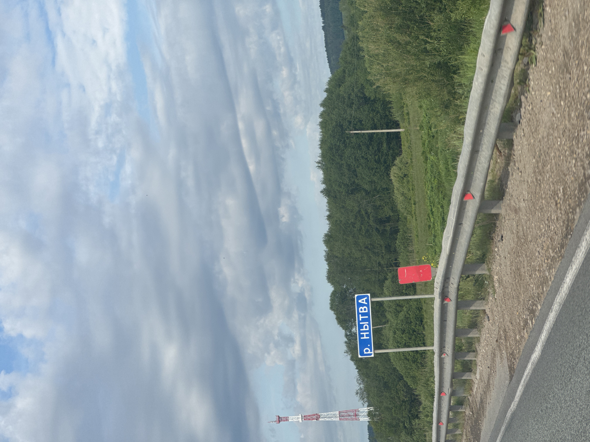 This image captures a daytime roadside scene, likely from a moving vehicle, along a highway in a rural, forested area in Russia, specifically near Шумиха. The sky, occupying the upper left portion, features a mix of bright white and grey clouds with patches of blue visible.
The scene includes a grey asphalt road on the lower right, bordered by a substantial metal guardrail equipped with multiple red triangular reflectors. Adjacent to the guardrail, the road shoulder is gravelly. Beyond the guardrail, a dense expanse of green trees and lush vegetation forms a forest that stretches across the middle ground and recedes into the distance, suggesting rolling hills or elevated terrain.
A prominent blue road sign with white Cyrillic text is visible on the right side, positioned behind the guardrail. The text reads "р. НЫТВА", which translates to "r. NYTVA", indicating a river or locality named Nytva. To the right of this sign, a red rectangular object, possibly another sign or marker, is partially visible. In the distant left, a tall, red and white striped lattice tower, resembling a communication or transmission mast, stands against the sky.
The weather appears partly cloudy but bright, indicating mid-day. The overall impression is that of traveling through a picturesque natural landscape typical of the Russian countryside.