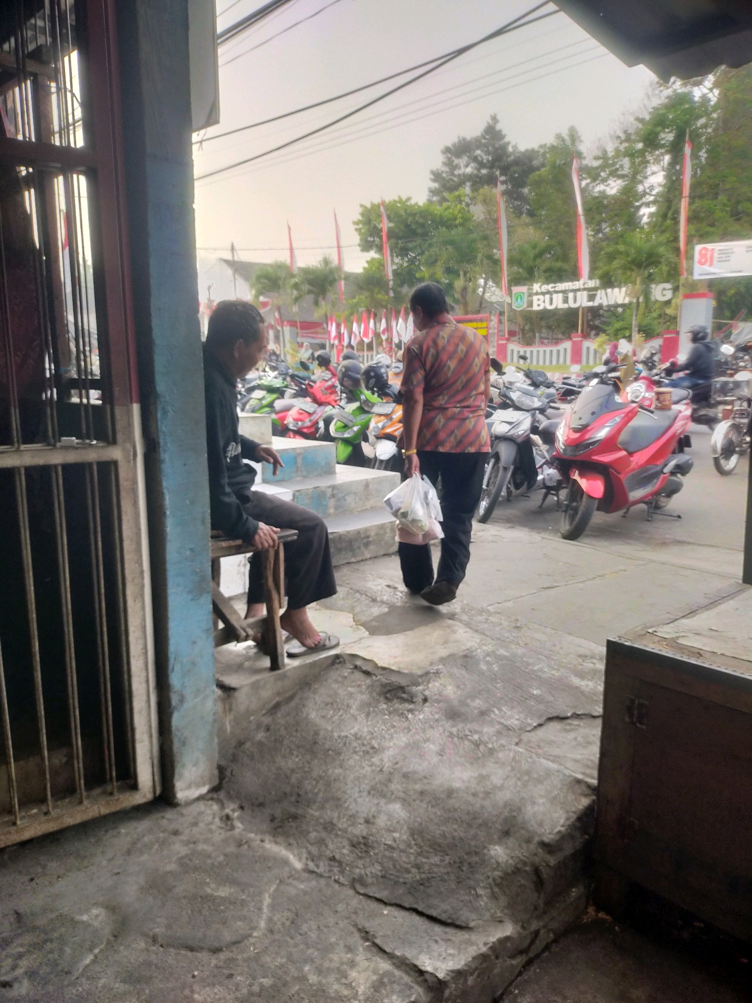 The image captures a street scene in Bululawang, Indonesia, with a prominent sign that reads "Kecamatan BULULAWANG". In the foreground, a man is seated on a stool by a gate. Further back, another man in a patterned shirt walks away from the camera, carrying plastic bags. The street is lined with parked motorcycles of various colors, including a red scooter in the center. Red and white flags are visible in the background, suggesting a local event or celebration. The overall environment appears to be an outdoor commercial area.