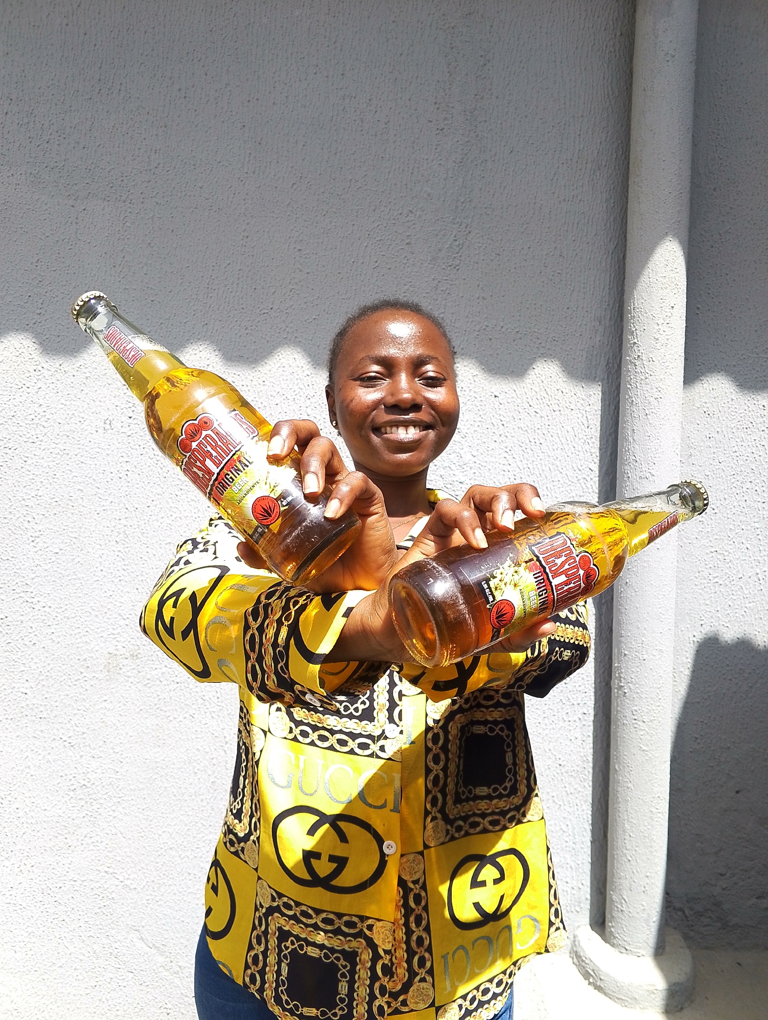 A young woman, likely in her early twenties, stands outdoors against a textured gray wall. She is holding two bottles of Desperados beer, one in each hand, angled upwards. She is wearing a vibrant yellow shirt with a black and gold patterned design featuring interlocking chains and the Gucci logo. Her facial expression is a wide, joyful smile, and her eyes are bright. The lighting suggests it is daytime, with shadows cast on the wall from an unseen source. The background appears to be a residential area or an open public space within the city of Jos, Nigeria, as indicated by the architectural style of the wall and the typical sunlight conditions.
