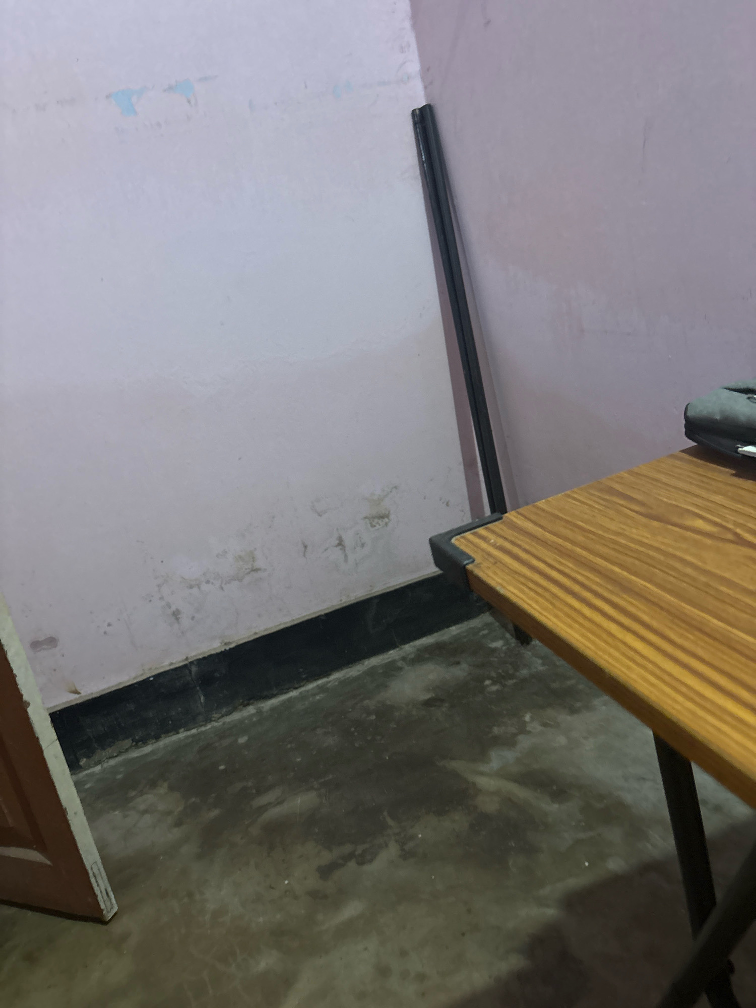 The image depicts an interior scene, possibly a room, in Jorhat, India. A wooden-topped table with dark metal legs is positioned in the foreground, occupying the right side of the frame. A dark object, possibly a bag or electronic device, rests on the table. To the left of the table, a portion of a wooden door is visible. The walls appear to be painted in a light pink hue, with some signs of wear and discoloration, particularly near the base of the wall. A dark skirting board runs along the bottom of the wall. A tall, dark object, possibly a thin pole or frame, leans against the wall in the background. The floor appears to be concrete and shows some water stains and a somewhat rough texture. The overall impression is of a simple, functional interior space.