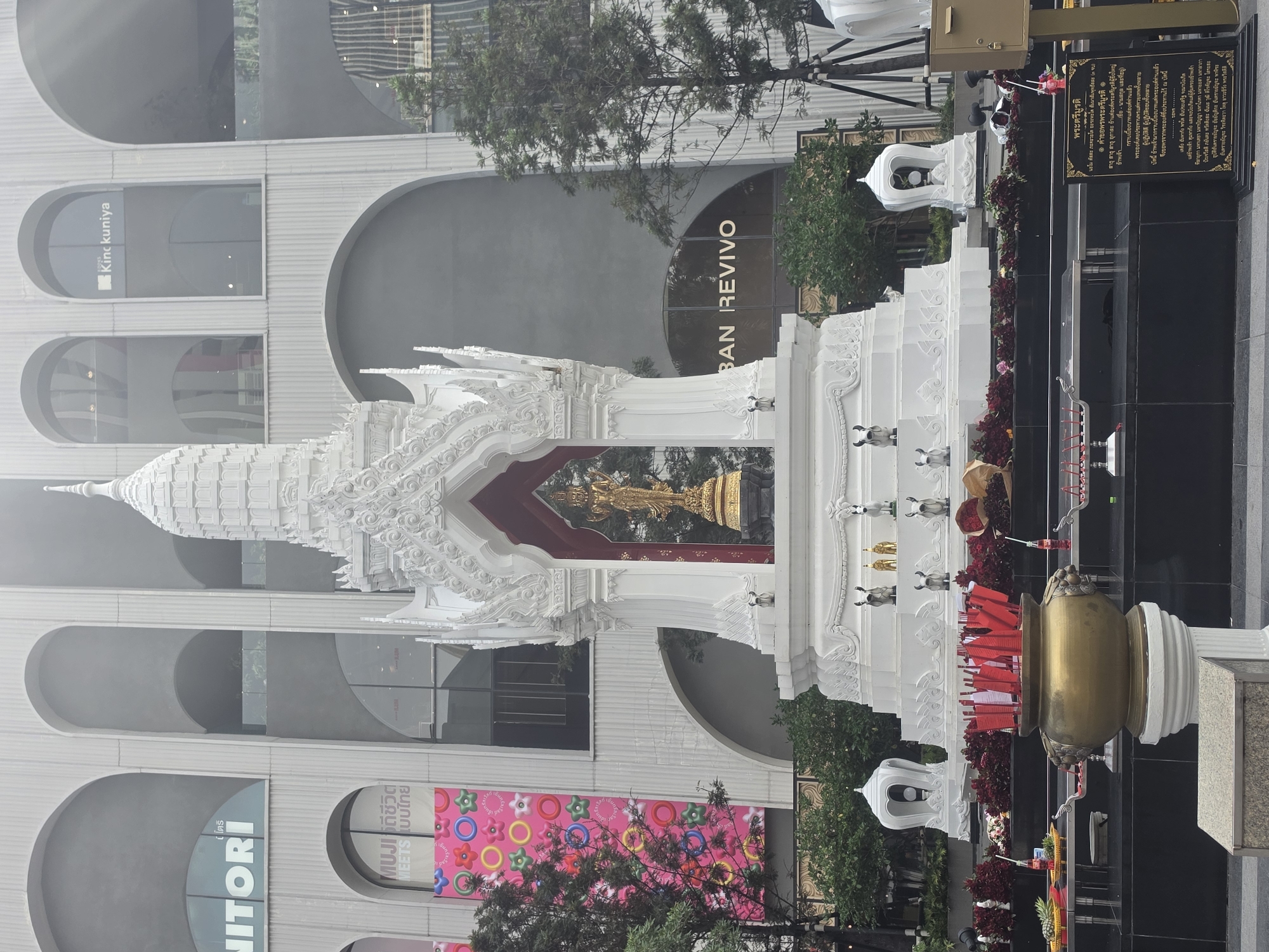 This outdoor scene in Bangkok, Thailand, focuses on a white Thai-style spirit house housing a golden statue. The ornate structure is located in front of a multi-story modern building with arched windows. Banners and signs for businesses, including "NITORI", "Kinokuniya" and "BAN REVIVO", are visible on the building's facade. The spirit house is adorned with offerings, flowers, and a metal pot filled with red incense sticks or flags. A black sign with Thai script stands next to the shrine. The atmosphere is slightly hazy, indicating possibly early morning or a humid day.