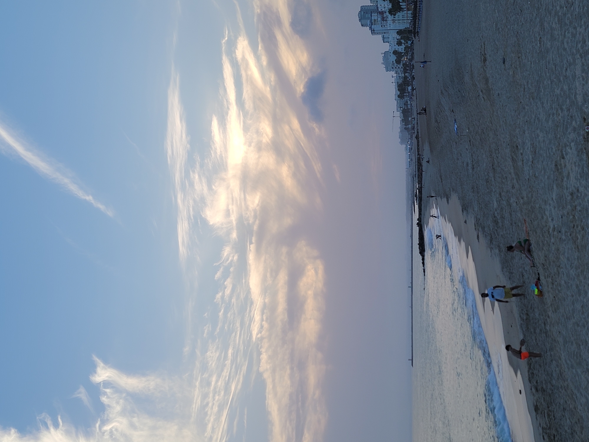 The image captures a serene evening on the beach in Quarteira, Portugal, with the soft hues of a twilight sky dominating the upper portion. Wispy clouds, tinged with the fading sunlight, stretch across the pale blue expanse. Below, the calm ocean gently laps against the sandy shore, its surface reflecting the muted colors of the sky.
On the beach, a few individuals are engaged in leisure activities. In the foreground, two young boys, one wearing an orange swim trunk, appear to be playing with a paddle game on the sand. Nearby, a man in a white t-shirt and yellow shorts stands, observing. Further back, a woman in a green swimsuit is seated, possibly resting. The beach extends towards a distant breakwater, marked by lighthouses, and the cityscape of Quarteira, characterized by white buildings and palm trees, lines the coastline. The overall atmosphere is one of peaceful relaxation as the day transitions into night.