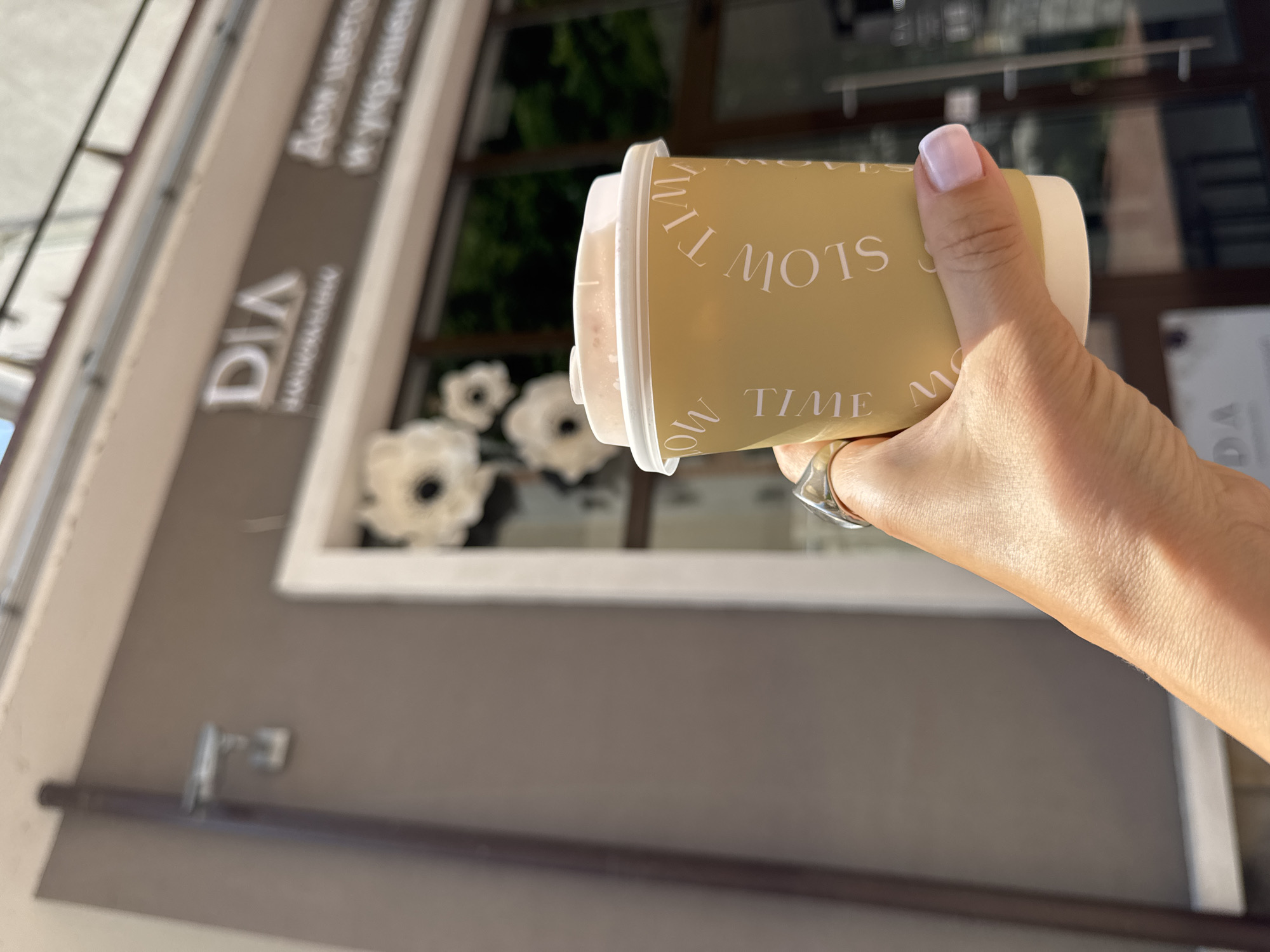 A close-up shot focuses on a hand holding a light brown paper coffee cup with a white lid. The cup has a repeating pattern of the words "SLOW TIME" in a light, almost white font. A silver ring is visible on the ring finger of the hand, and the fingernails are painted a light pink color.
In the blurred background, a building with a brown facade is visible. A sign above a storefront reads "DIA" in large, white, sans-serif letters, with smaller text below it. To the right of "DIA," there is more text in white, which appears to be Cyrillic. The storefront has large windows with white decorative elements that resemble flowers.
The image appears to be taken outdoors on a sunny day, as indicated by the bright lighting and shadows. The overall impression is that of a casual moment, possibly outside a cafe or shop in a city. While the specific location of Estosadok, Russia, is mentioned in the prompt, there are no explicit visual cues within the image itself that definitively confirm this location.