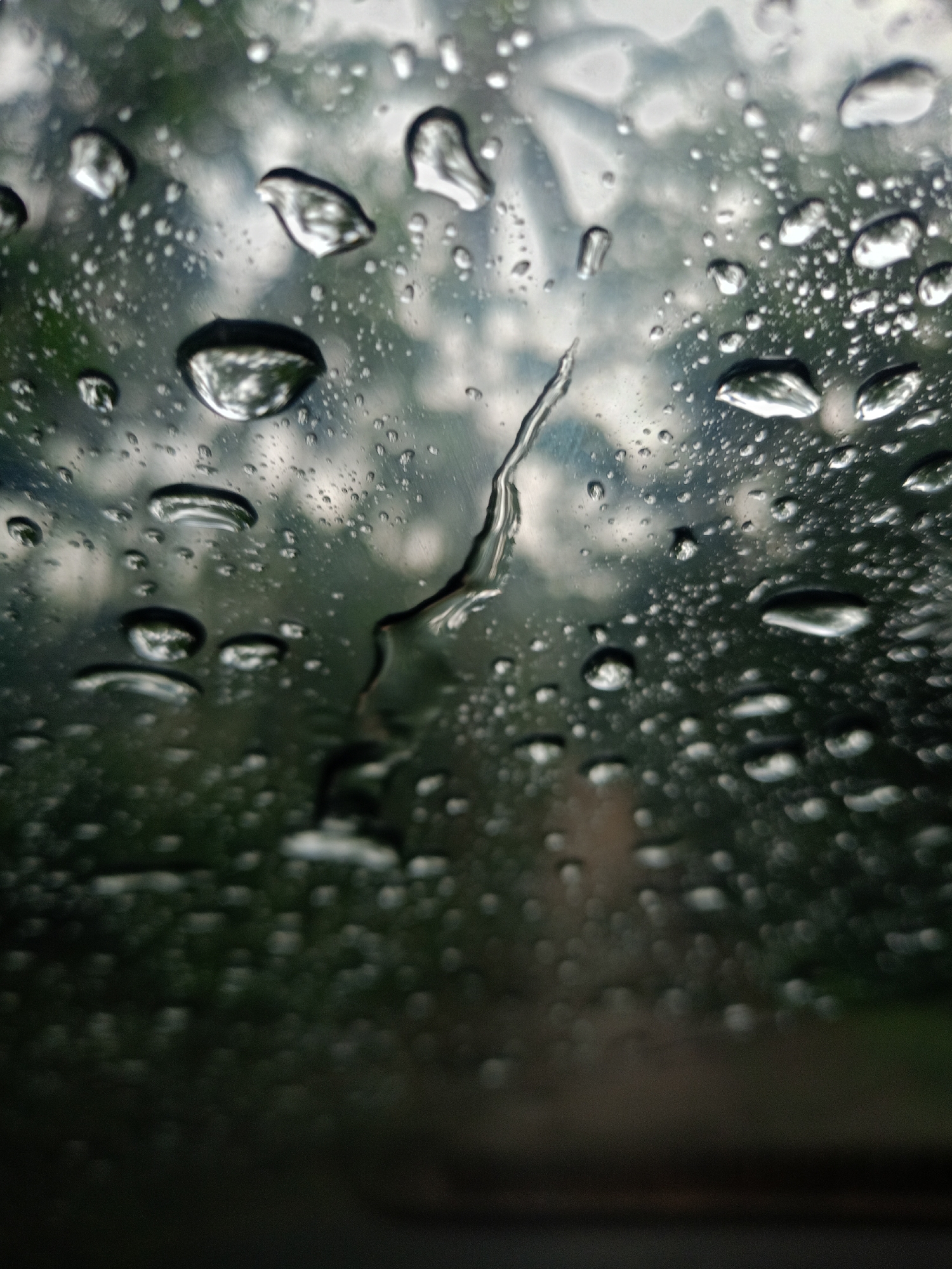 The image shows a close-up view of raindrops on a glass surface, likely a window. Through the water droplets, an obscured view of a cloudy sky and green foliage (possibly trees) can be seen. A streak of water runs vertically down the center of the glass. The foreground is dark and out of focus. The location is unknown.