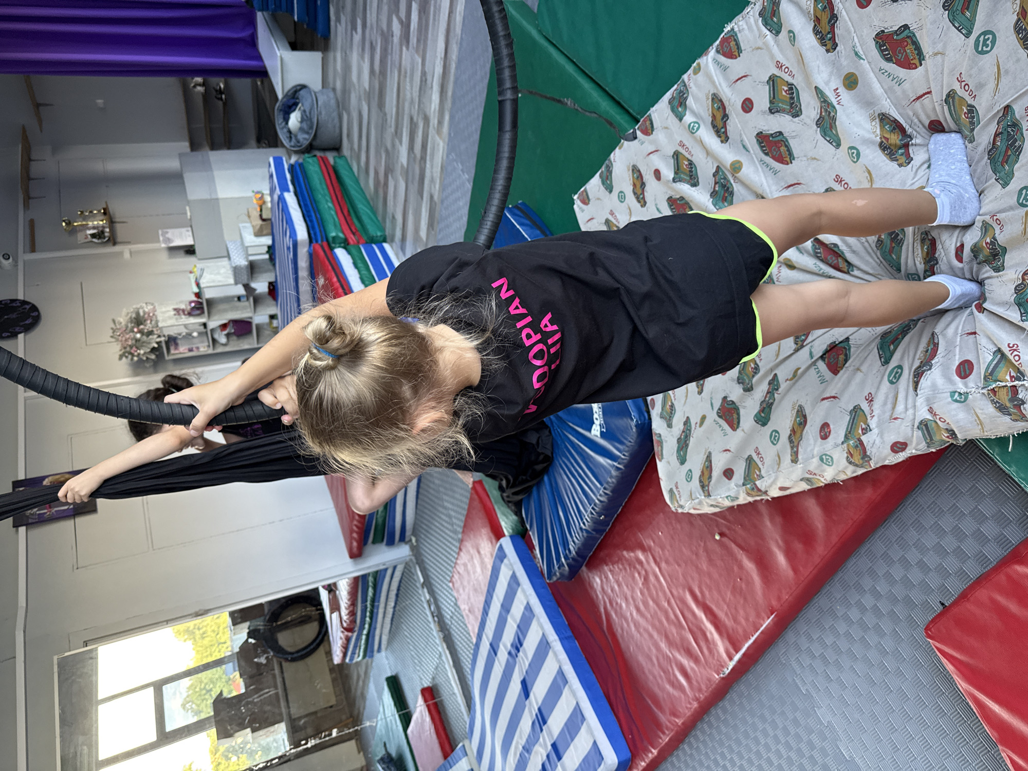 This is a photograph taken at a gym or indoor playground in Dnipro, Ukraine. The image features a young girl with blonde hair tied in a bun, wearing a black t-shirt with pink lettering that reads "BOOK DOPIAN A". She is suspended in the air, holding onto a black aerial hoop or fabric apparatus. Her legs are extended, and she is wearing white socks. In the background, there are various pieces of gymnastic equipment, including colorful mats stacked on the floor, a purple curtain, and shelves with trophies. The scene suggests an aerial or gymnastic training session. The lighting indicates it is daytime.