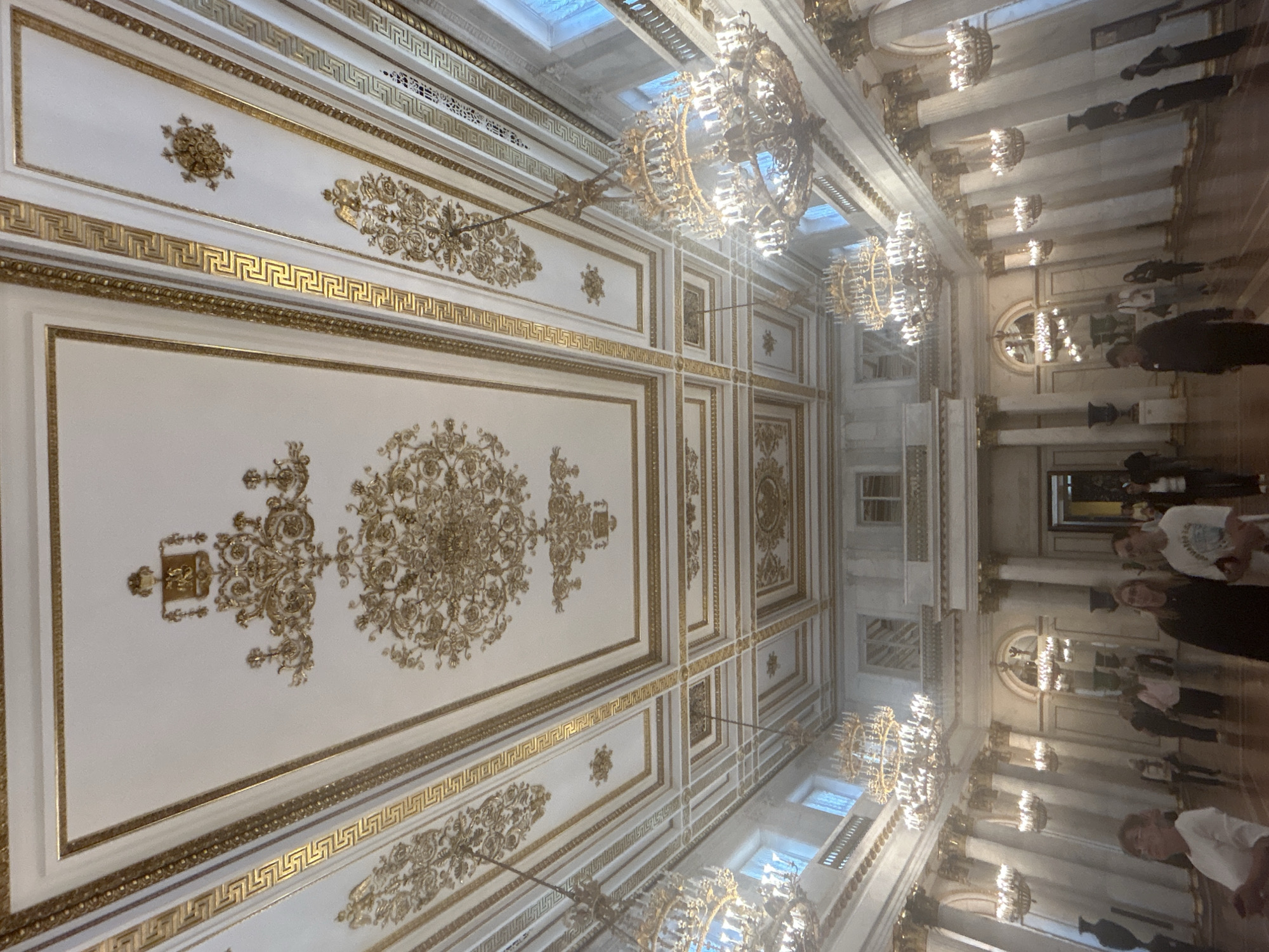 The image depicts an ornate interior of a grand hall, likely a museum or palace, with a highly decorative ceiling and multiple chandeliers. The ceiling is white with intricate gold embellishments and Greek key patterns along the borders of recessed panels. Large, multi-tiered chandeliers with many lights hang from the ceiling. The hall has tall white columns with gold capitals, and the walls are light-colored, featuring arched doorways and windows. Several people are visible, walking and standing in the hall, appearing to be tourists or visitors. The floor is a polished wooden surface that reflects the light. The overall impression is one of opulence and historical grandeur.