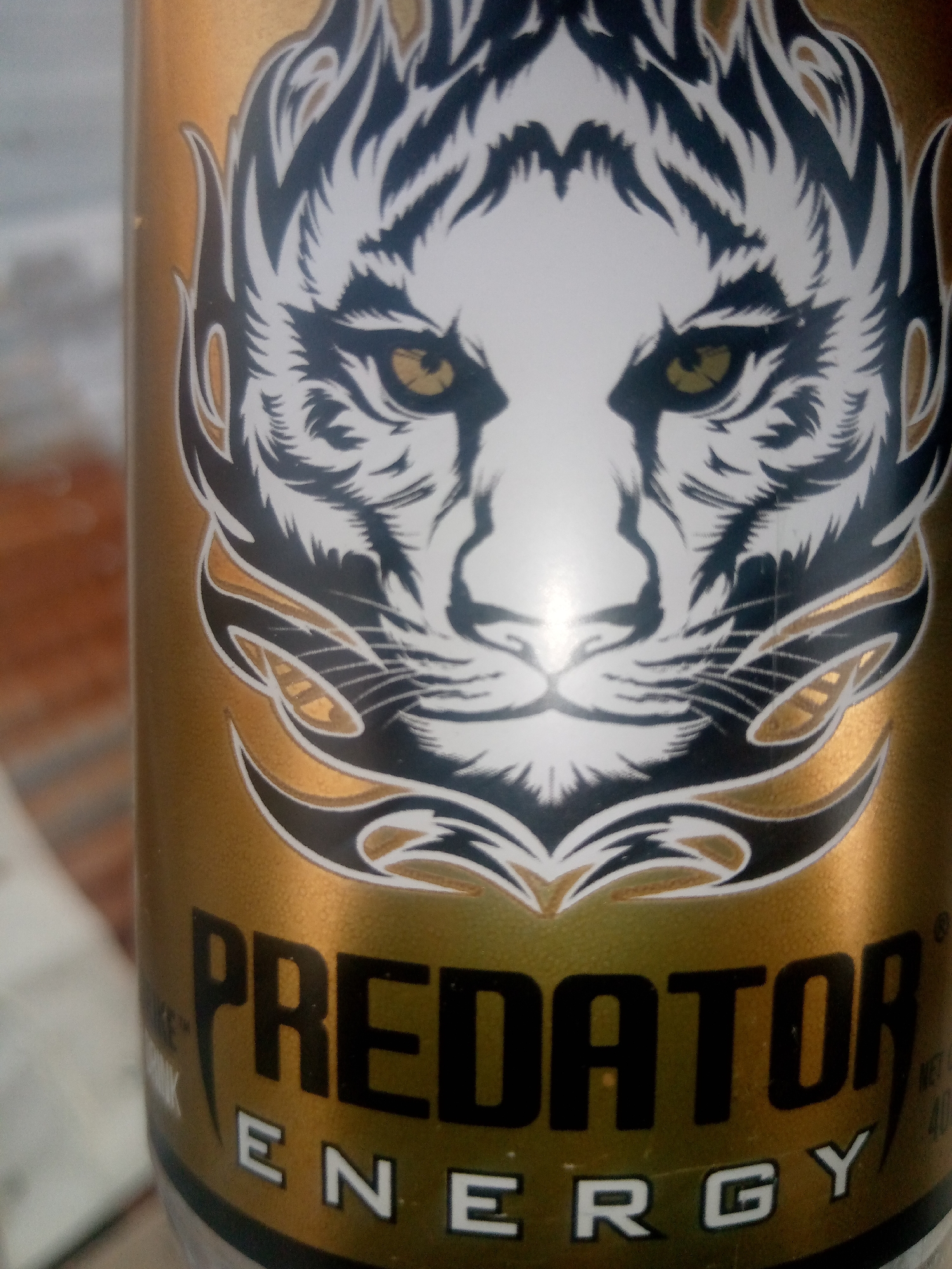 The image shows a close-up of a gold-colored can of "PREDATOR ENERGY" drink. The can features a graphic of a white tiger's face with black stripes and yellow eyes, framed by black and gold tribal-like designs. The text "PREDATOR ENERGY" is prominently displayed in large, black, sans-serif font. The background is out of focus and appears to be a wooden surface. The image does not depict any specific location in Jalingo, Nigeria, or any people.