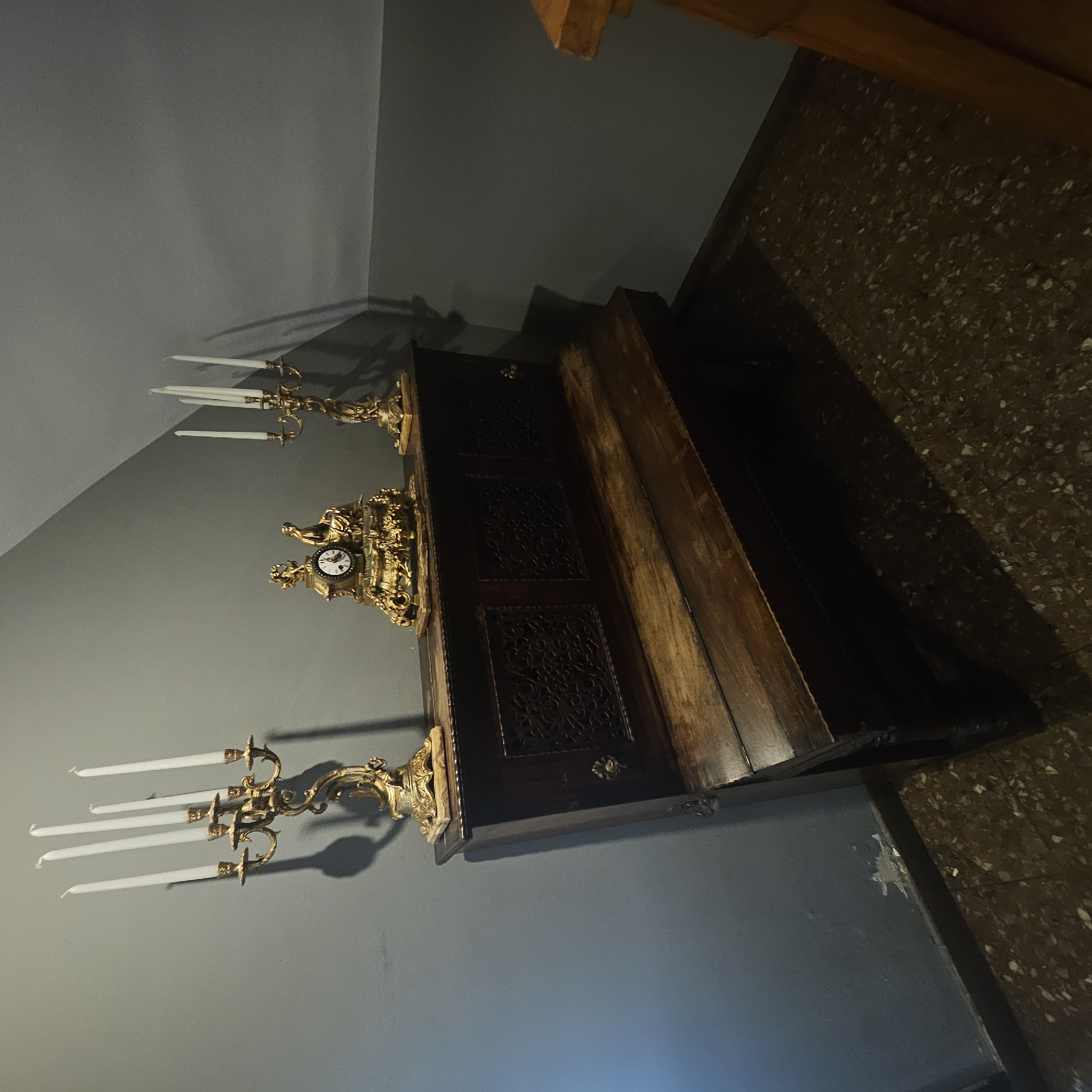 In Almadrones, Spain, a dark wooden bench with intricate carvings sits against a gray wall. On top of the bench, a golden mantel clock with figurines is displayed between two ornate golden candelabras, each holding several white candles. The floor is a speckled mosaic pattern.