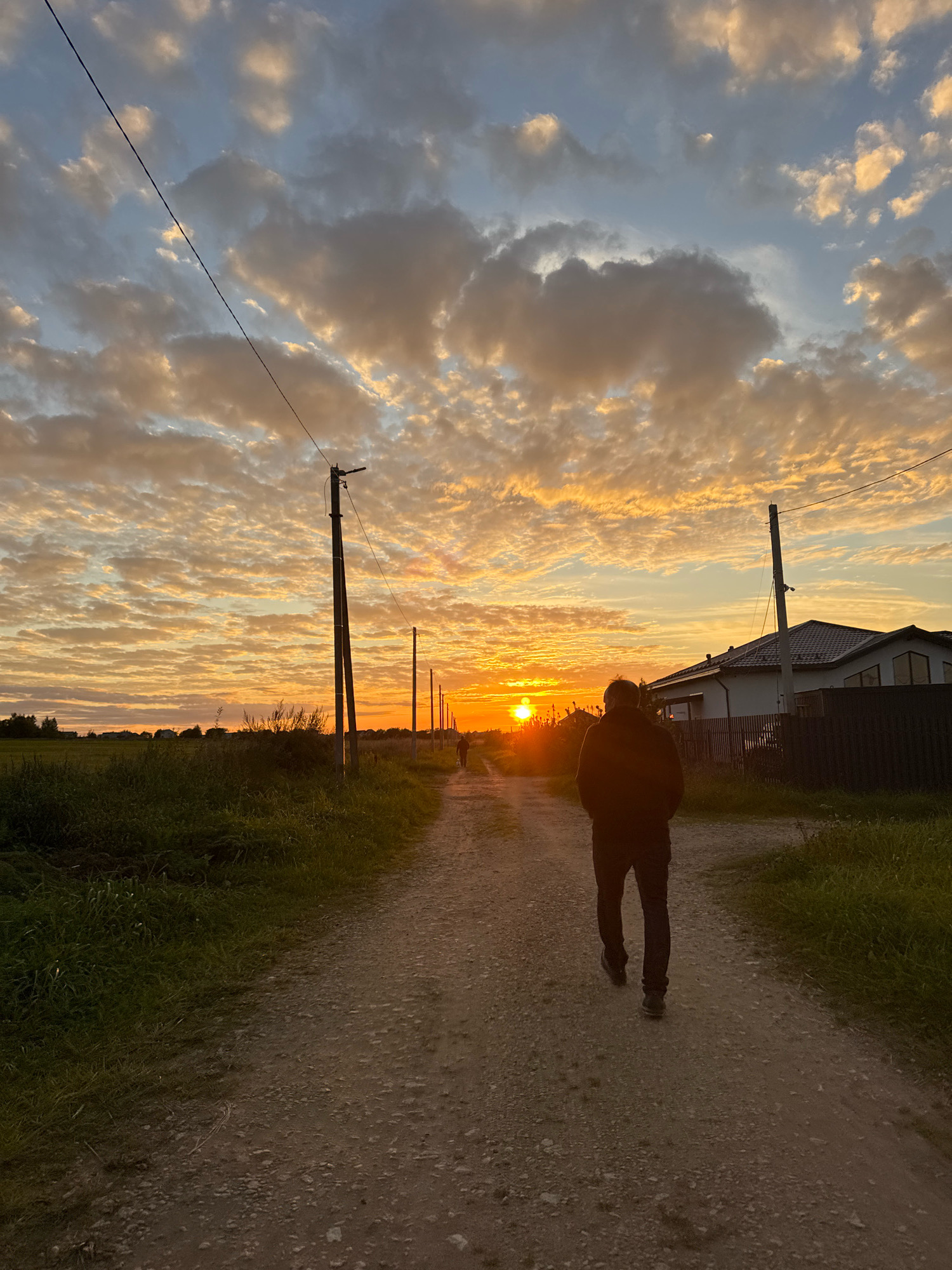 A vertical, full-shot photograph captures a sunset scene in the village of Kempelevo, Russia. In the foreground, a person wearing dark clothing walks away from the camera down a gravel road. The road is lined with green grass on both sides. In the middle ground, several utility poles with wires stretch across the scene, and a white house with a dark roof is visible on the right. In the background, the sun sets behind a tree line, casting a warm orange glow across the sky, which is filled with scattered clouds. The overall impression is one of a serene, rural evening.