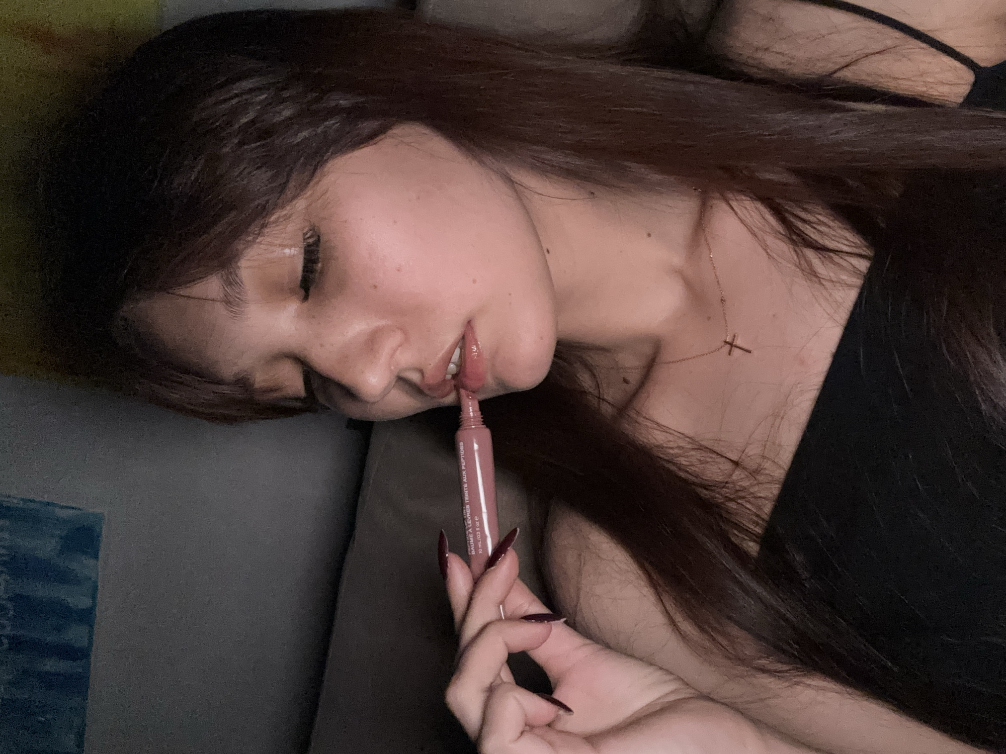 In a dimly lit indoor setting, a young woman with long, dark hair and a black top is applying lip gloss. Her eyes are closed, and she holds a tube of lip gloss with her left hand. A delicate cross necklace is visible around her neck. The background appears to be a couch with a textured blanket. No specific location in Novokuznetsk, Russia can be identified.