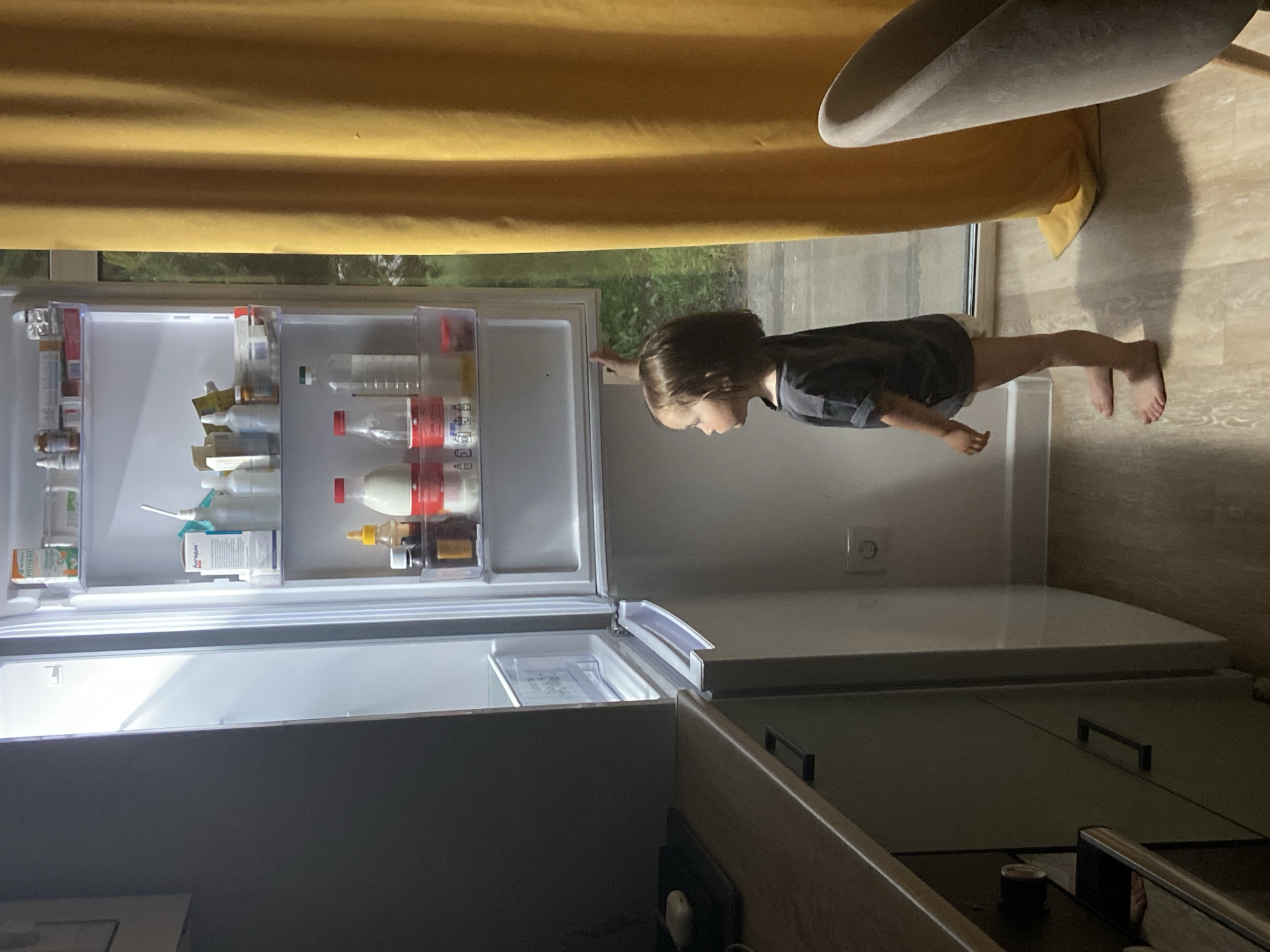 A child is standing near an open refrigerator in what appears to be a kitchen. The refrigerator is stocked with various food and beverage items. The kitchen features wooden cabinets, a gray chair, and a yellow curtain. The image appears to be taken indoors, and the exact location within Tyumen, Russia, cannot be determined from the available visual information.