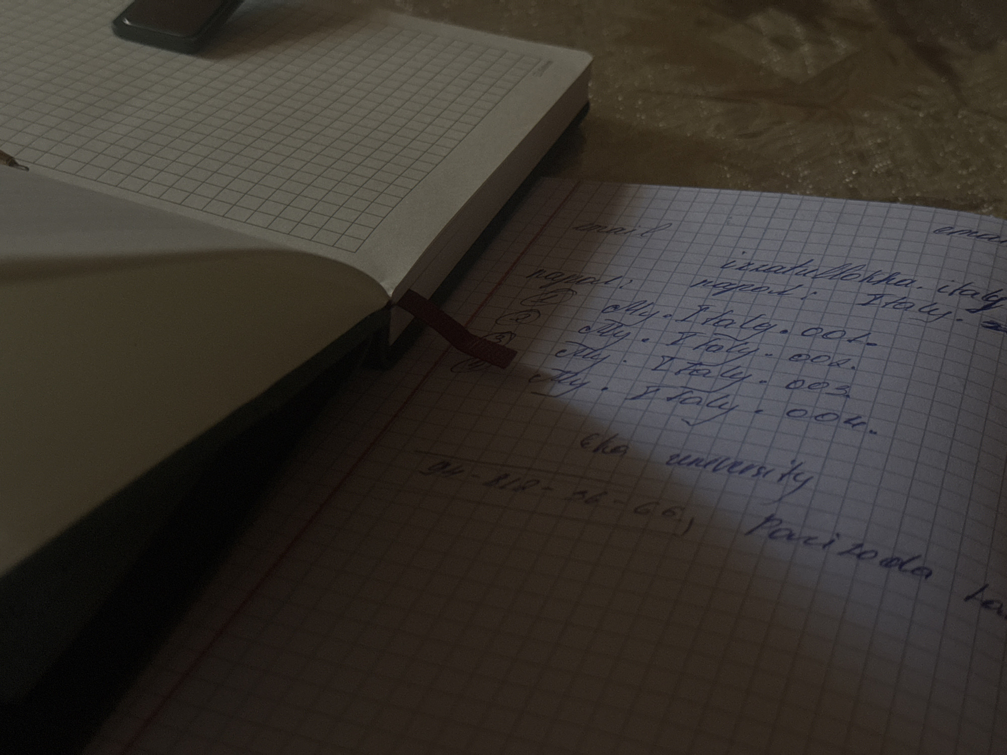 The image shows a close-up view of two open notebooks on a textured surface. The notebook on the left has a grid pattern on its pages and a red ribbon marker. A dark, rectangular object, possibly a phone, is partially visible on the top left page of this notebook. The notebook on the right, also with a grid pattern, is filled with handwritten text in blue ink. Visible words and phrases include "email", "napoli", "Italy", "city. Italy. 001.", "city. Italy. 002.", "city. Italy. 003.", "city. Italy. 004.", "the university", and a sequence of numbers and letters: "94-812-26-66, Pari 20 ola". The lighting suggests the photo was taken in a dimly lit environment. There is no information in the image that indicates a location in Navbahor, Uzbekistan.