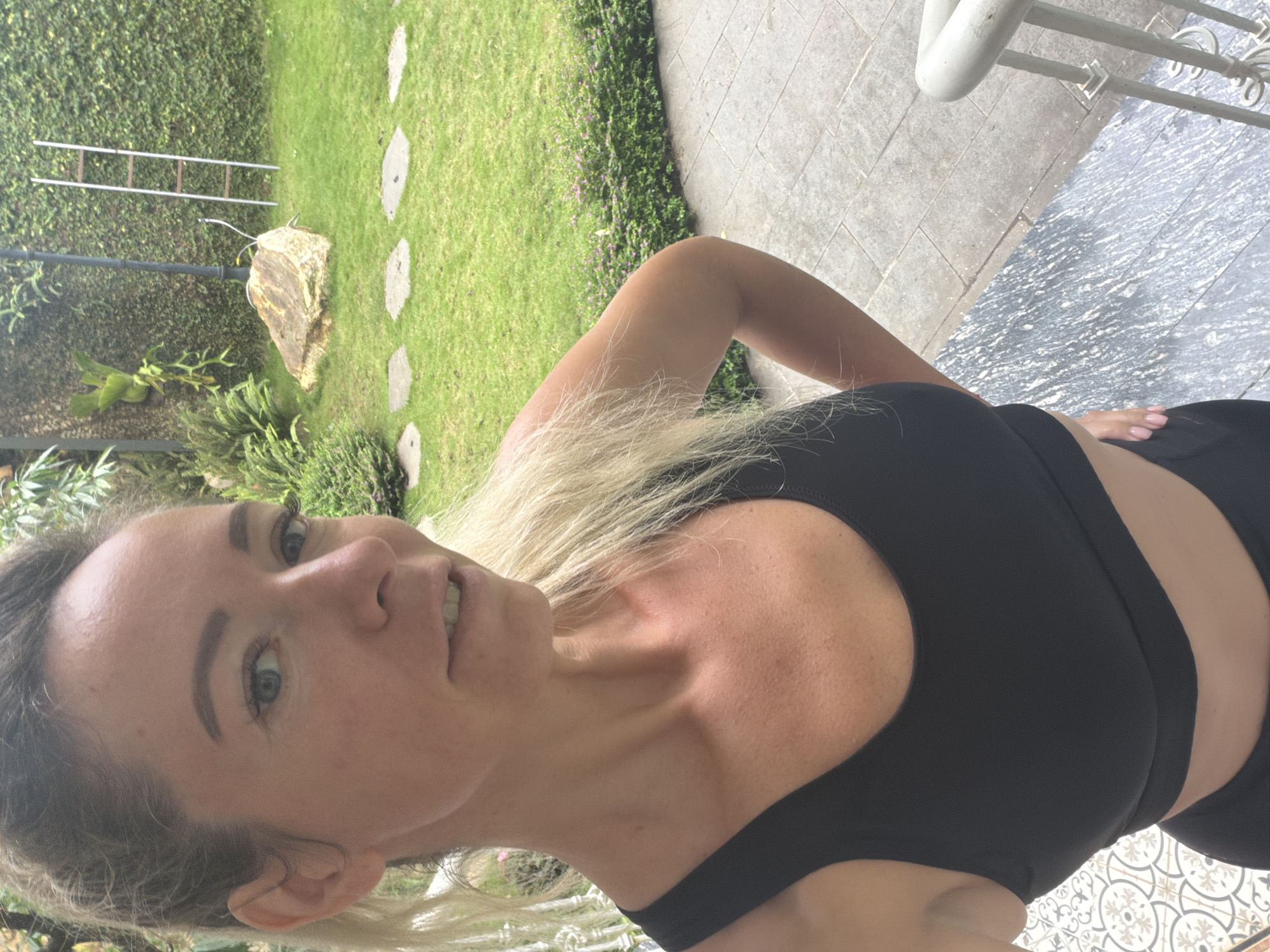 A blonde woman in a black sports bra and black leggings stands on a tiled patio, taking a selfie. She is positioned in the foreground, with a lush green garden visible in the background. The garden features a large rock, stepping stones, manicured hedges, and a vine-covered wall. A silver ladder leans against the wall. The scene appears to be outdoors in a residential or resort setting. The location is identified as Xuân Trường - Đà Lạt, Vietnam.
