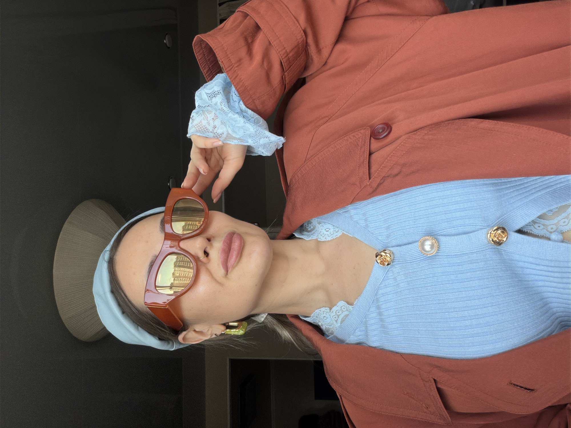A close-up, rotated portrait image features an individual, likely a woman, posing for a selfie indoors. The person has light skin, plump lips, and dark hair partially covered by a light blue, fabric headband. They wear large, rectangular, brown-framed sunglasses with highly reflective golden lenses, which show the reflection of an architectural cityscape, suggesting an outdoor urban environment.
The individual is dressed in a light blue, ribbed knit top with delicate lace trim along the neckline and cuffs. The top is adorned with decorative gold buttons, some designed as roses and one featuring a pearl center. Over this, a rust-colored or terracotta-toned jacket or coat is worn, visible through its structured sleeve and main body, with visible stitching and a matching button. A square, gold-toned earring is visible on the left ear, and a small gold nose ring is present. The person's hand, with manicured light-colored fingernails, is casually raised near their face, touching the sunglasses.
The background is dark and somewhat indistinct, with a tall, light-colored, textured object (possibly a lamp or art piece) partially visible on the left. The overall impression is of a stylish, candid moment captured during the daytime, likely in Krasnoyarsk, Russia, as suggested by the location context. No visible text is present in the image.