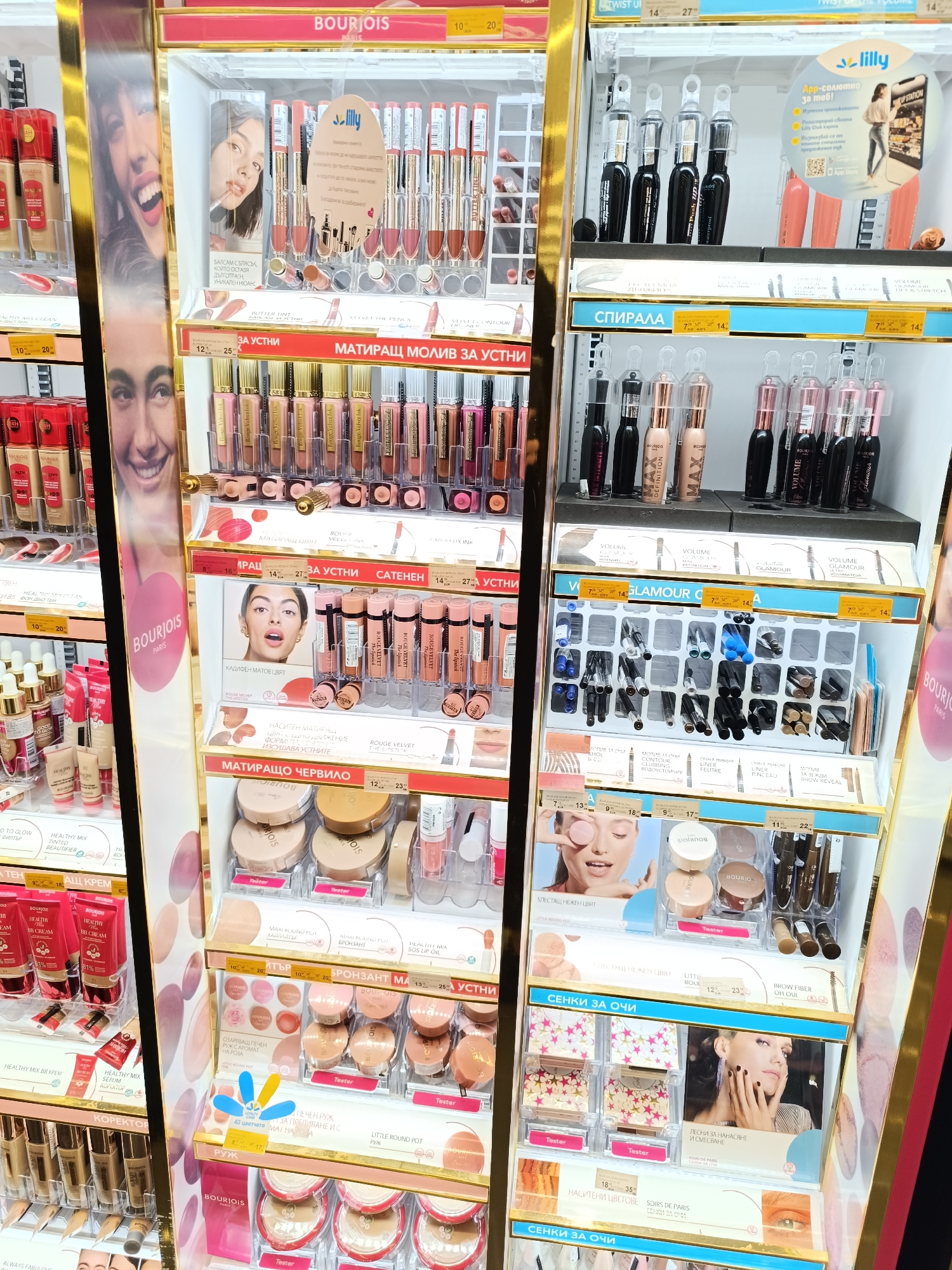 The image shows a cosmetics display, likely inside a retail store in Plovdiv, Bulgaria. The brands Bourjois and Lilly are prominently featured. The shelves are filled with various makeup products including lipsticks, lip liners, mascaras, eye shadows, blushes, and BB creams. Prices are displayed in Euros. The products are well-organized, with testers available for some items. There are advertisements and promotional materials integrated within the display. The environment appears to be well-lit and clean.